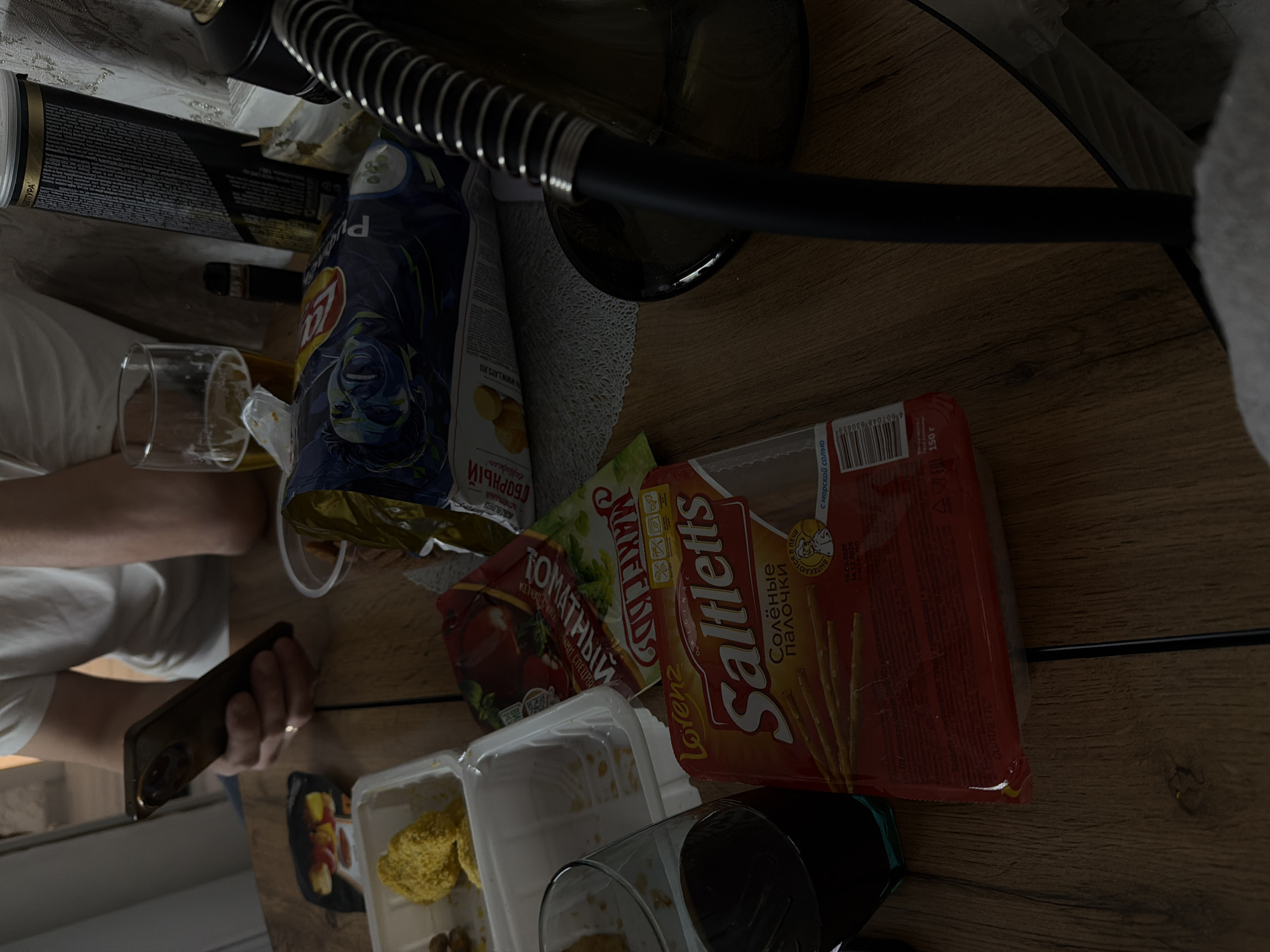 The image shows a cluttered table with various food items and objects. A person's arm and hand are visible, holding a smartphone. The table is covered with items such as a package of Lay's chips, a pack of Lorenz Saltletts pretzel sticks, a container of salad, and a food container. The environment suggests a casual setting, possibly indoors.