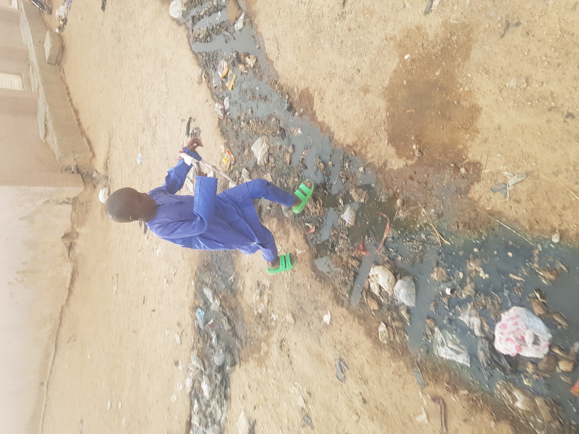 A young person in a blue garment and green sandals is walking near a sewage trench filled with debris in Dutsinma B, Nigeria. The surrounding area appears to be a dusty, unpaved street with a building visible in the background.