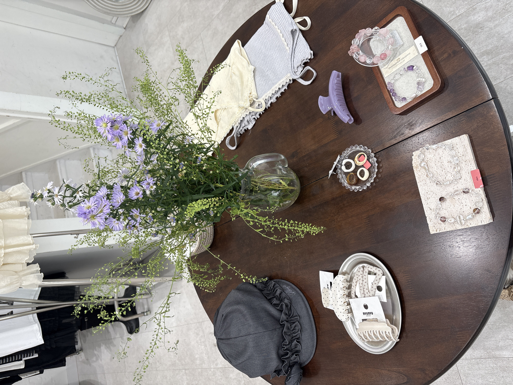 An overhead, slightly angled shot captures a well-arranged display table in what appears to be a retail boutique, likely located in London, United Kingdom. The central subject is a round, dark brown wooden table, showcasing a variety of fashion accessories and delicate apparel.
In the center of the table, a clear glass vase holds a vibrant bouquet of purple and white aster-like flowers mixed with slender green foliage, providing a natural focal point. To the top left, two delicate camisole tops or bralettes, one cream and one light blue, both featuring lace trim, are neatly laid out. Adjacent to them is a large, solid lavender claw hair clip.
Moving clockwise, a rectangular wooden tray with a felt lining displays two beaded bracelets, one in pink and clear tones, and another in purple and clear. A price tag reading "$9.9" is visible, along with a white card that possibly states "HAPPINESS HAPPENING." Further around, a small, scalloped clear glass dish contains several small, chunky rings or earrings in white and brown shades.
To the right, a rectangular off-white stone block serves as a display for another clear beaded bracelet, accompanied by a pink tag with the number "5293." Towards the bottom right, a silver oval tray holds various hair accessories, including two patterned claw clips (one white with black polka dots, one cream) and a floral hair tie. Visible branding on the tags includes "NYUNYU" and "NRUJUAT." Completing the display at the bottom left is a dark gray cloche-style hat with a ruffled brim, featuring faint embroidered text that appears to be "Madara."
In the background, out of focus, clothes racks laden with white and black garments suggest a clothing store setting. The overall presentation is clean and organized, indicating an inviting retail environment designed for showcasing merchandise. No people or specific activities beyond the static display are depicted.