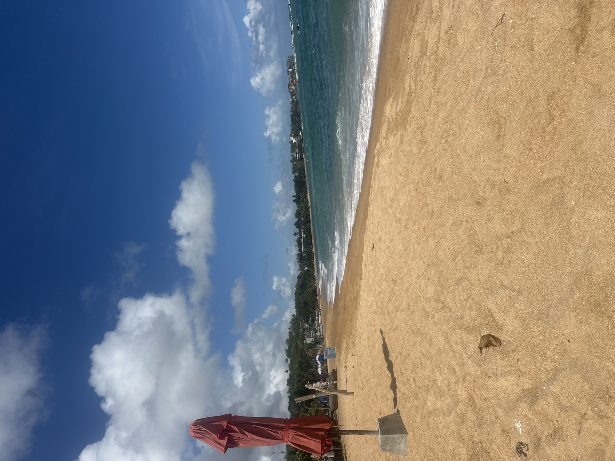 The image shows a beach scene in Rumassala, Sri Lanka. The sky is blue with scattered white clouds. The ocean is turquoise with white waves. The beach is sandy with a reddish-brown hue. A closed red beach umbrella stands on the sand. A few dry leaves are scattered on the sand. The edge of the beach is lined with trees and some buildings.