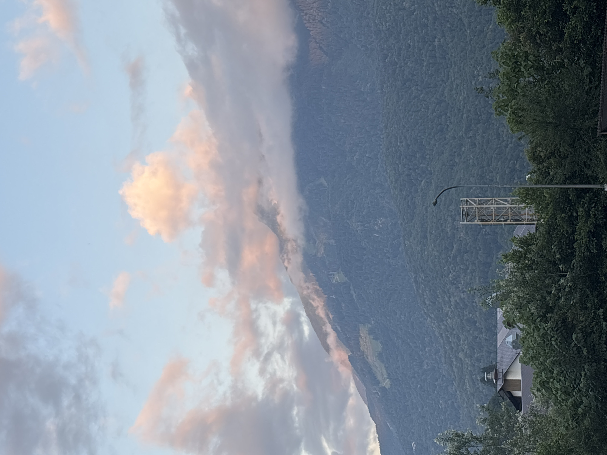 The image shows a mountainous landscape under a sky with pinkish clouds. In the foreground, there are green trees and some buildings, including a structure that appears to be a ski lift station or a similar industrial facility. The mountains are covered in dense forest, with some ski slopes visible on one of the higher peaks. The overall scene depicts a natural setting with some human infrastructure, likely in a mountainous resort area. This is consistent with the environment of Krasnaya Polyana, Russia, known for its ski resorts and mountainous terrain.