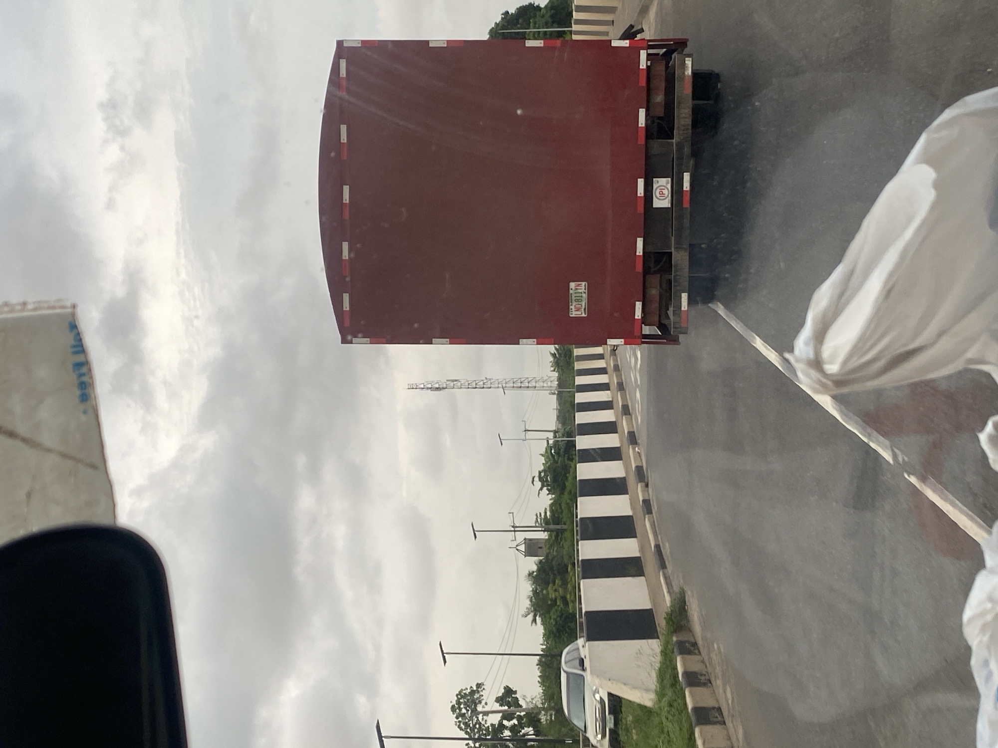 This image depicts a rear view of a large, dark red truck driving on a road in Ikorodu, Nigeria. The truck has a license plate that reads "LND 811YN". The road is flanked by a black and white striped barrier and a paved shoulder. In the background, there are trees, streetlights, and a tall cell tower. The sky is overcast with clouds. The image appears to have been taken from another vehicle, as evidenced by the reflection on the windshield and a white plastic bag in the foreground.