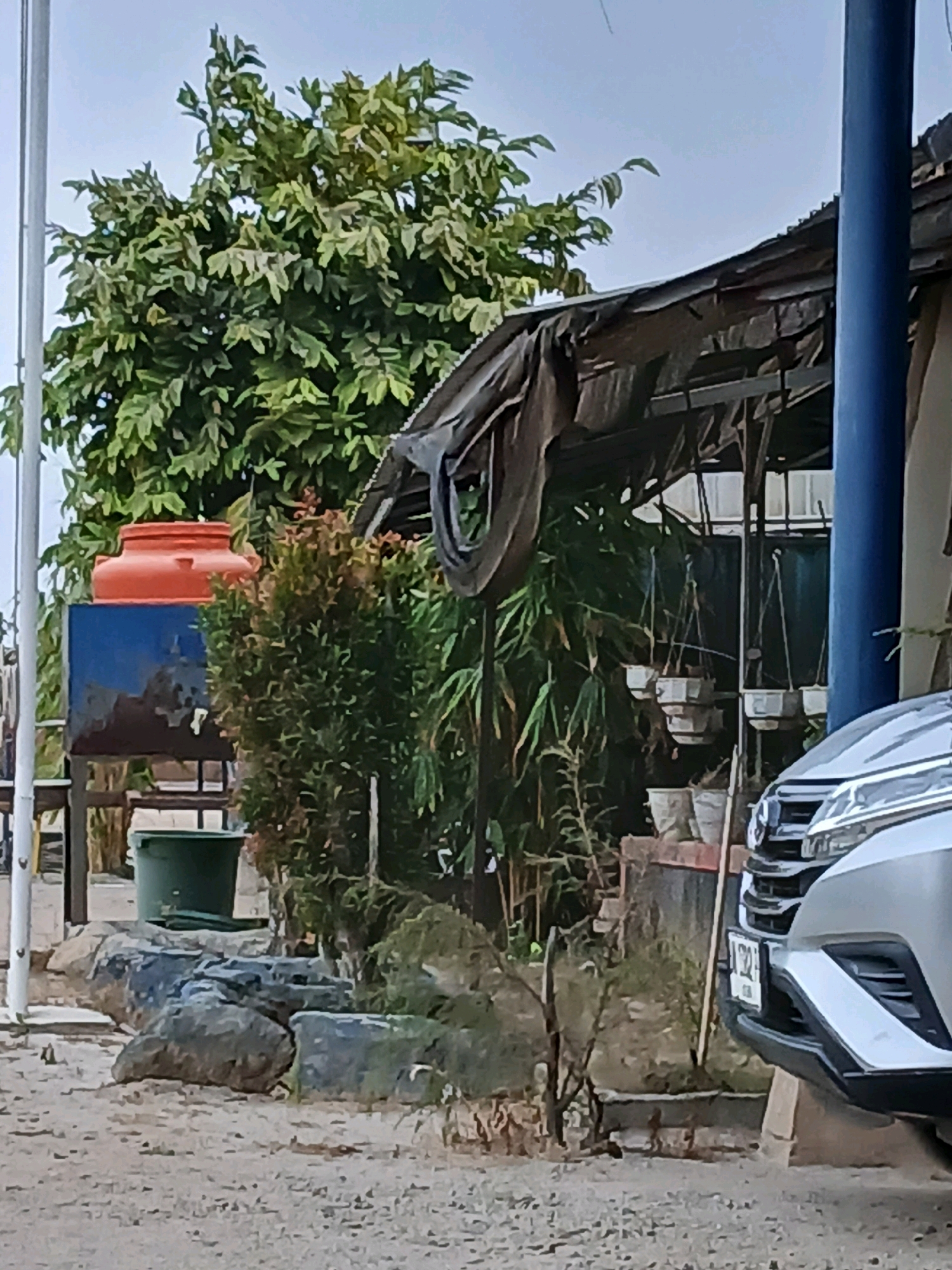 A silver car is parked in front of a structure with a corrugated metal roof and a dark tarp covering part of it. The car's front bumper and headlight are visible. Behind the car and structure, there is dense green foliage from trees and bushes. In the foreground, there is a sandy or gravelly ground with several large rocks. To the left of the structure, there is a large orange water tank on top of a blue structure, and a green bucket. A thin white pole is visible to the far left of the frame. The scene appears to be outdoors, possibly in a semi-rural or mixed-use area of Pangkalpinang, Indonesia, given the presence of a water tank and the style of the structure, which could be a home, a small business, or a workshop. The sky is overcast. There are no people visible in the image.