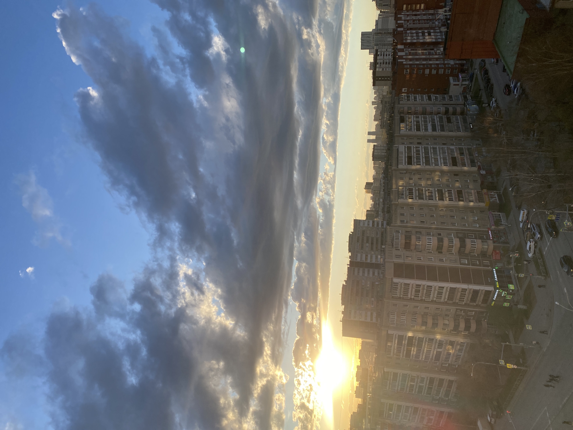 The image captures an urban landscape during either sunrise or sunset, with the sun low on the horizon. The sky is a prominent feature, dominated by large, elongated dark clouds interspersed with patches of clear blue sky. The sun, located towards the right, emits a strong golden glow, illuminating the edges of the clouds and casting a warm light over the scene. A green lens flare is visible in the clouds near the sun.
Below the horizon, a densely packed city skyline extends across the frame. Numerous multi-story buildings, likely residential or commercial, line the streets. These structures display varied architecture, with some featuring red brick facades and others appearing in lighter gray or beige tones. Many windows are visible across the building exteriors.
At street level, a road runs between the buildings, with several cars parked and a few in motion. Pedestrians can be observed on sidewalks and utilizing zebra crossings. Various traffic signs and road markings are discernible. Sparse, bare trees are scattered in the foreground, suggesting a late autumn or winter season. On one of the buildings, a green sign clearly displays the Cyrillic word "Аптека," which translates to "Pharmacy." The overall scene depicts a cloudy yet illuminated cityscape, characteristic of Yekaterinburg, Russia, as the day begins or ends.