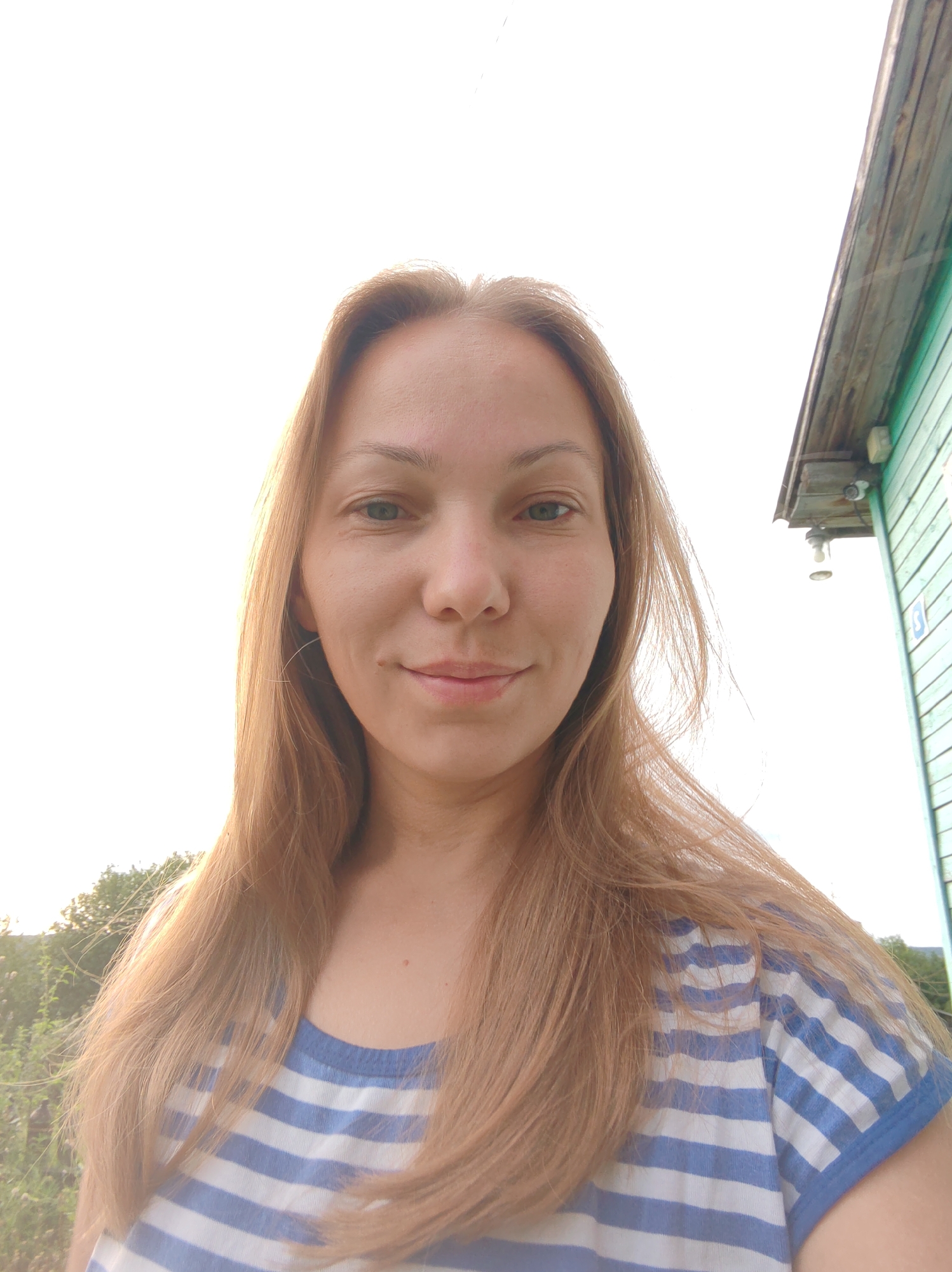 The image shows a medium shot of a woman outdoors, with a bright sky behind her and the side of a green wooden building on the right. The woman has long, light brown hair that is parted in the middle and falls to her shoulders. She is wearing a blue and white striped t-shirt. Her face is turned towards the camera, and she has a slight smile. Her skin appears fair, and she has green eyes.
The building on the right is painted green and appears to be made of horizontal wooden planks. There are a few outdoor lights attached to the side of the building, one of which is visible towards the top right of the image. A small portion of foliage is visible in the background to the left of the woman. The overall lighting suggests it is daytime, possibly late afternoon or early morning, given the softness of the light. There are no clear indicators of the specific location within Chukhlem, Russia, beyond it being an outdoor, possibly rural or suburban, setting.