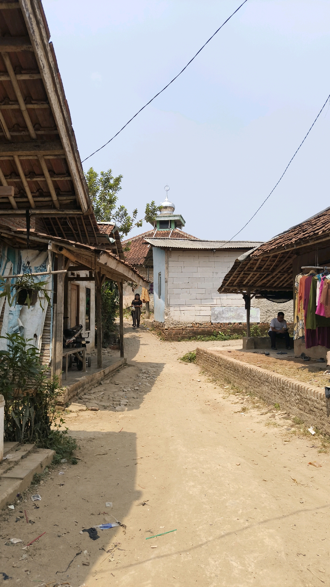 The image depicts a sunlit, unpaved village street in Tenjo, Indonesia, bordered by simple, low-rise structures. On the left, a building with a wooden beam and tile roof overhangs the path, featuring a white sheet draped on its exterior and a hanging plant. Further down the path, a person is walking, silhouetted against the light. To the right, a brick wall lines the street, beyond which is a covered area with clothes hanging on a rack. A man is seated under this shelter, facing away from the camera. In the background, a mosque with a green-domed roof and a minaret is visible, surrounded by other buildings with tiled roofs. The ground is dusty and littered with small debris. The sky is clear and bright.