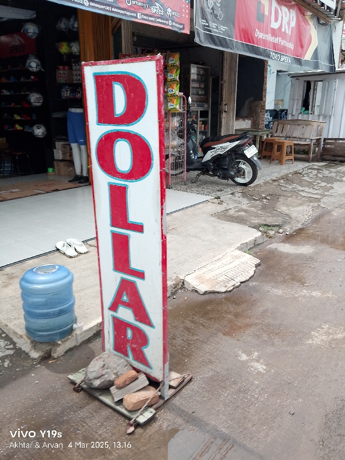 The image shows a signpost with "DOLLAR" written in red letters on a white background. The signpost is on a street corner in Jatibarang, Indonesia. The location is identified by the coordinates -6.468 latitude and 108.302 longitude. There is a blue water cooler on the left side of the signpost and a broken concrete block on the ground. A motorcycle is parked in front of a shop across the street. There is a bench and some other objects in front of the shop. The street is made of asphalt and there is a puddle of water on the ground.