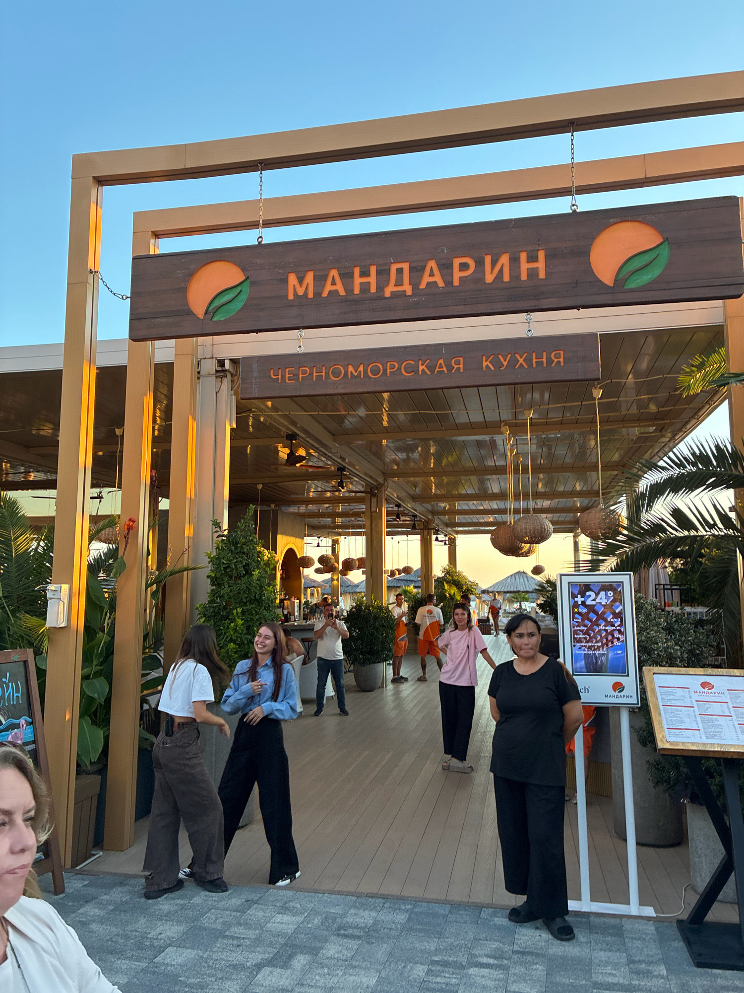 The image shows a sunny day in Sochi, Russia, outside a restaurant named "Мандарин" (Mandarin). The restaurant's entrance is decorated with a wooden sign featuring an orange graphic and Cyrillic text. The sign is suspended by chains from a wooden structure. Below the main sign, another wooden sign reads "ЧЕРНОМОРСКАЯ КУХНЯ" (Black Sea Cuisine).
Several people are visible. In the foreground, a woman in a black outfit stands facing the camera, looking slightly to her left. Behind her, two young women are engaged in conversation, with one laughing. Another woman in a pink shirt and dark pants stands near them. Further back, a man in an orange uniform is walking away from the camera, and other people are seen inside the restaurant, including a man taking a photo.
The environment is a walkway with wooden flooring, surrounded by greenery and plants. The restaurant appears to be open-air, with a roof supported by wooden beams and columns. Decorative pendant lights hang from the ceiling. A digital sign displaying the temperature (+24°) and other information is placed near the entrance. The sky is clear and blue. The overall impression is that of a lively, casual dining establishment in a warm climate.