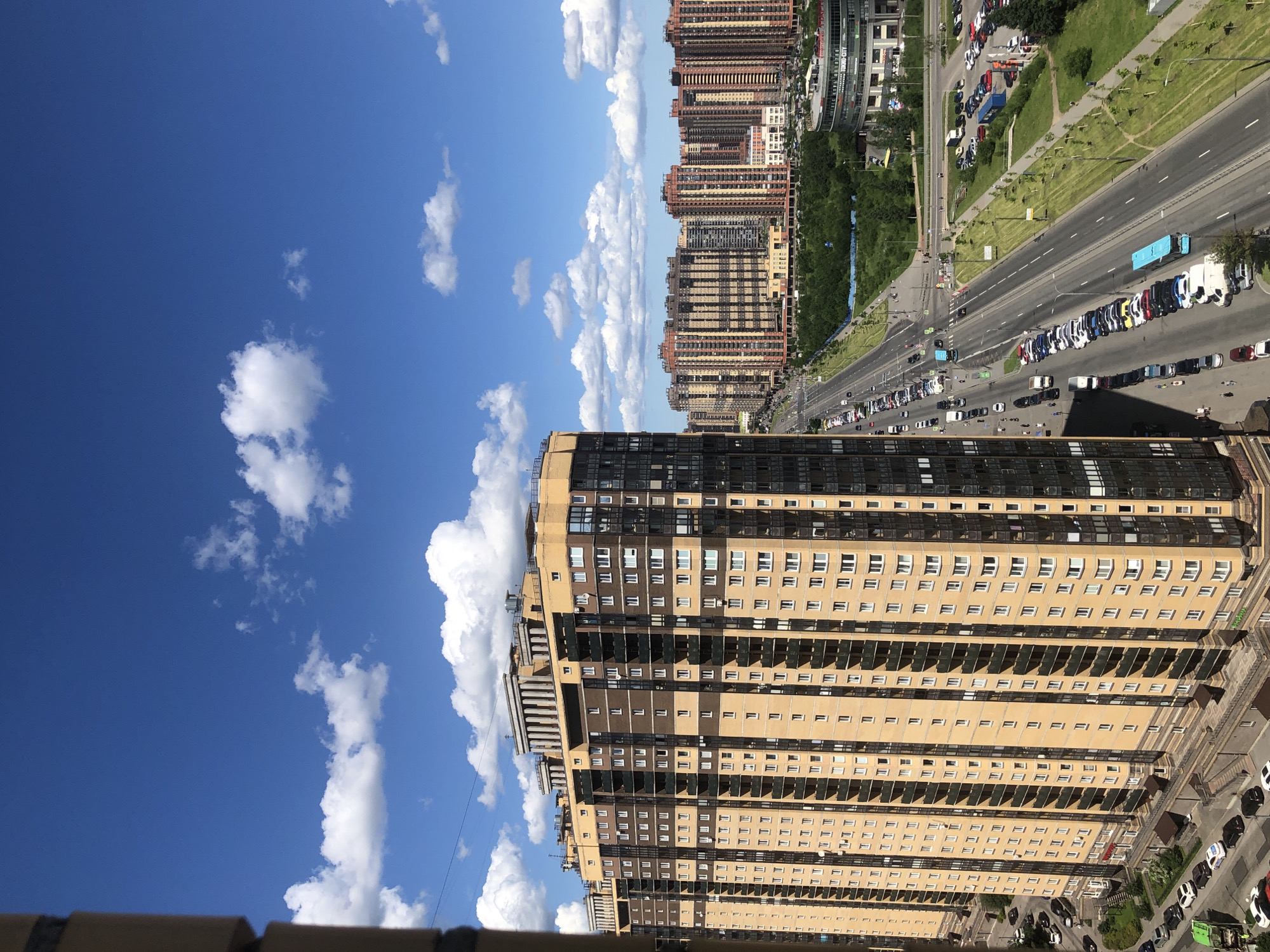 An elevated, eye-level, wide shot captures a bright, sunny day in a modern urban environment. The sky is a vibrant blue, dotted with fluffy white clouds. In the foreground, a tall, multi-story residential building dominates the left side of the frame, its facade a combination of beige brick and dark glass. Its many balconies create a repeating pattern of light and shadow.
To the right of the building, a wide, multi-lane road stretches into the distance, lined with parked cars. Several blue buses are visible among the traffic. The road is bordered by green spaces, including manicured lawns and trees. Further in the distance, a cluster of similarly tall residential buildings, characterized by their colorful, patterned facades, form a dense cityscape. One building in the mid-ground features a circular, modern architectural design with signage visible. The overall impression is one of a developing, contemporary city district under a clear, pleasant sky.