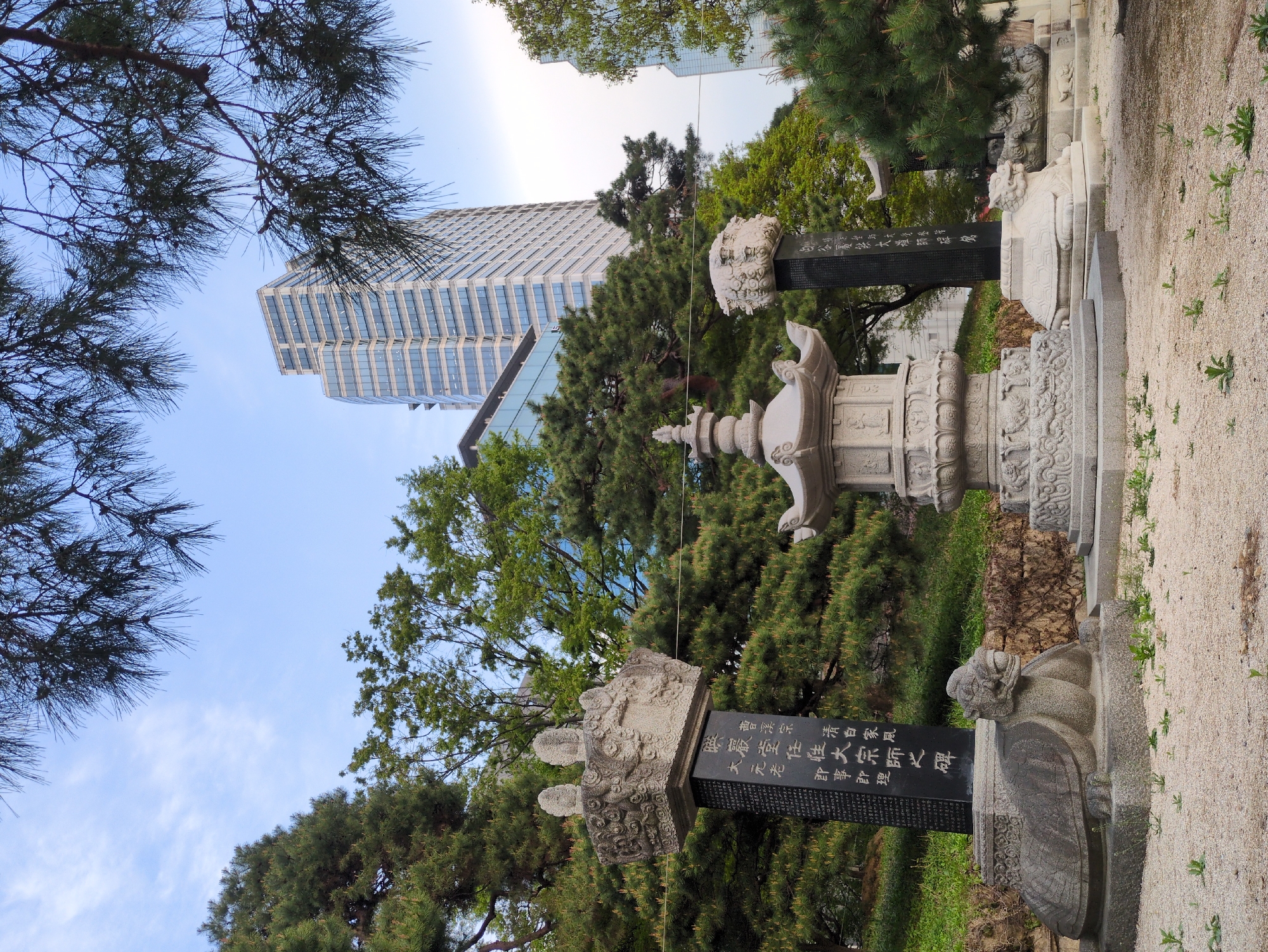 The image shows a traditional Korean garden in Seoul, South Korea, with ancient stone monuments standing amidst trees and a modern skyscraper in the background. The monuments include carved stone pillars and decorative elements, some with inscriptions in Chinese characters. The garden features gravel pathways and lush greenery, contrasting with the tall, glass-clad building visible in the distance, suggesting a blend of historical and contemporary aspects of the city. The sky is visible above the trees, and the photo is taken from a slightly low angle, emphasizing the height of the structures.