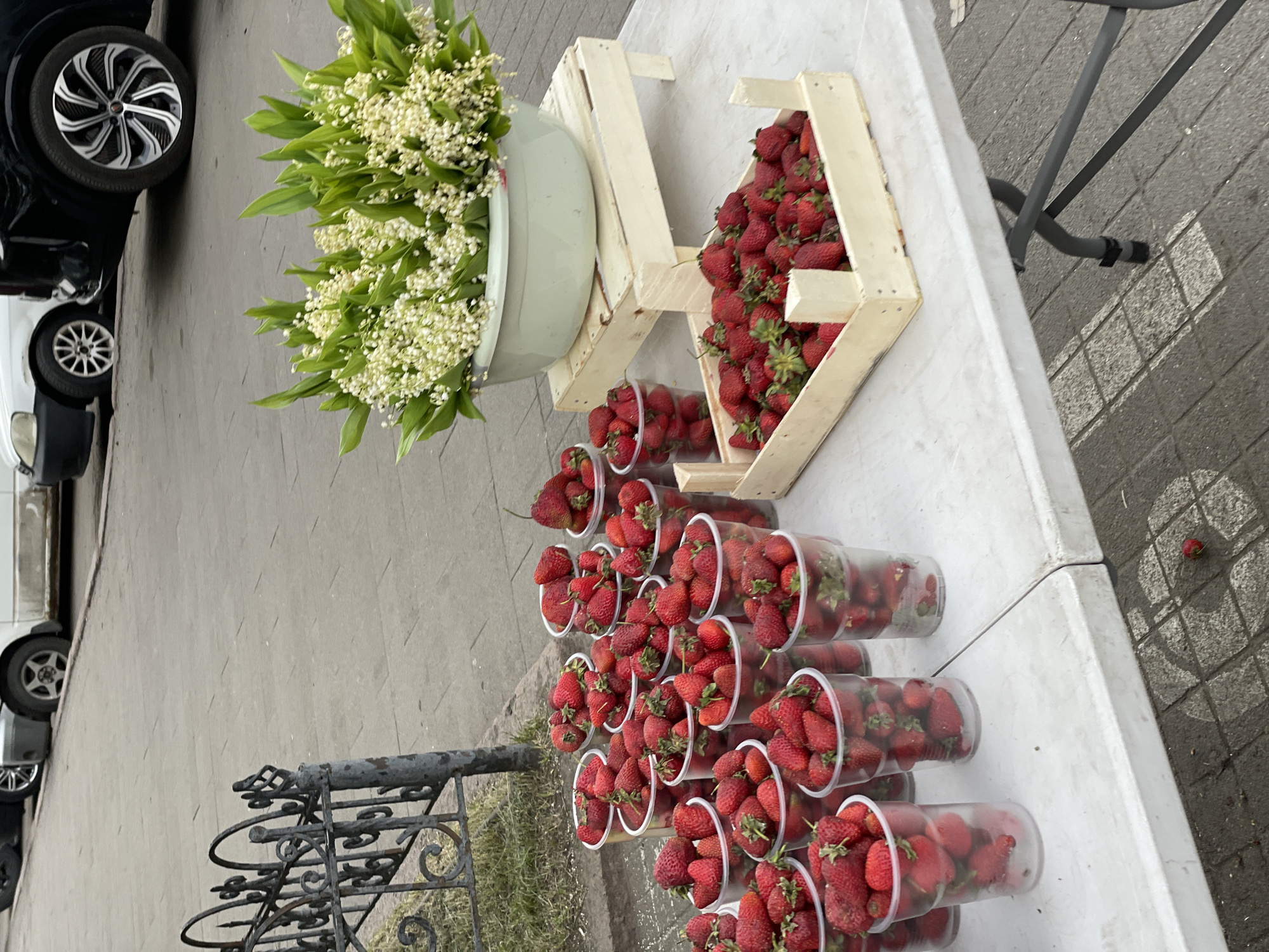 On a paved city sidewalk, next to a decorative metal fence with intricate scrollwork, a white folding table displays a variety of fresh produce. Prominently featured are numerous clear plastic cups filled with bright red strawberries, arranged in multiple rows. To the right of the cups, a light-colored wooden crate overflows with more strawberries. A large, pale green basin filled with a dense bouquet of white lily-of-the-valley flowers sits elevated on a small wooden platform behind the strawberries. In the background, cars are parked along the street. One fallen strawberry is visible on the paving stones in the foreground. The scene suggests an outdoor market stall likely located in Krasnoyarsk, Russia, given the context.