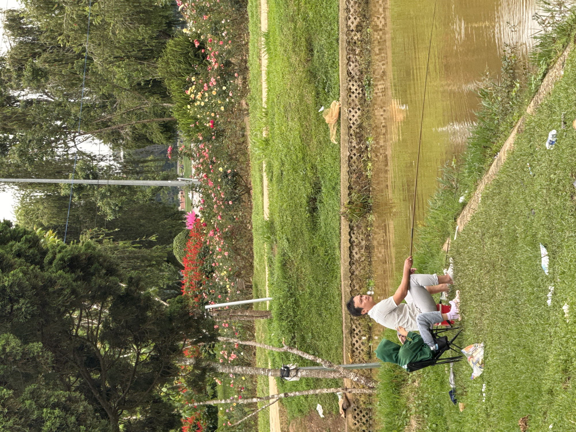 A man and child fishing in Phường Xuân Hương - Đà Lạt, Vietnam. The man, wearing a striped shirt and light grey pants, holds a fishing rod, the line cast into the muddy water. A child wearing a green hoodie sits in a black camping chair beside him. The area is grassy, with litter scattered around. A variety of trees and flowering plants are in the background.