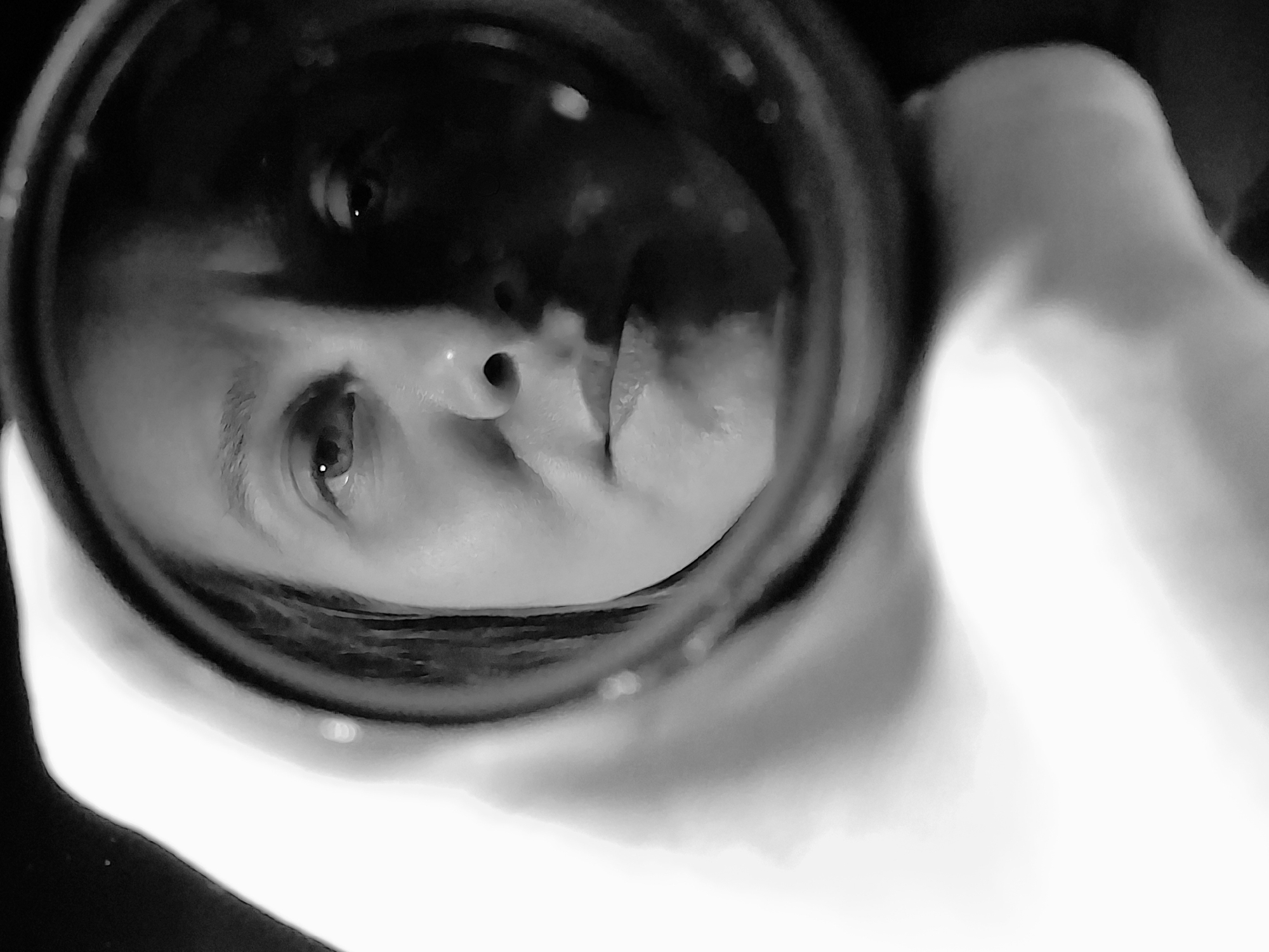 The image is a black and white, close-up shot of a face reflected in a circular object, possibly a lens or a small mirror. The face appears to be that of a person with light skin. The nose, one eye, and part of the mouth are clearly visible. The surrounding area within the circular object is dark with some lighter patches, potentially representing the background or the reflection's imperfections. The object is being held by a hand, partially visible in the blurred foreground. The image is taken at an angle, adding a dynamic element to the composition. There is no indication of the location being in the city of Chaikovsky, Russia.
