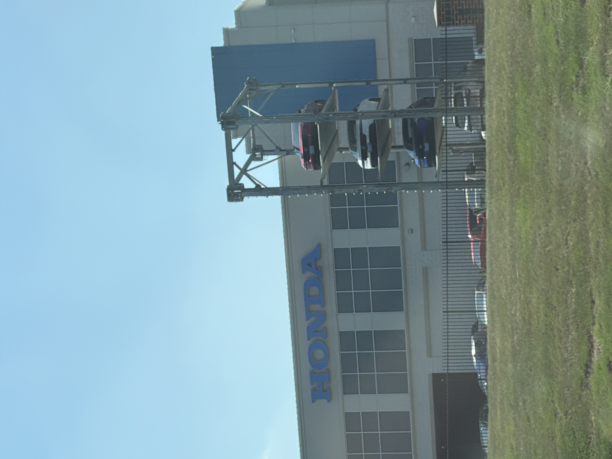 This is a wide, slightly blurred outdoor photograph of a Honda dealership. The building has a light-colored facade with a large blue panel on the right side. The word "HONDA" is displayed in large blue letters above a row of windows. To the right of the building, a car elevator holds three cars stacked vertically: a red car on top, a white car in the middle, and a dark blue car on the bottom. In front of the building, a black metal fence encloses a grassy area with several cars visible behind it. The sky is clear and blue. The location is a Honda dealership in Largo, United States of America.