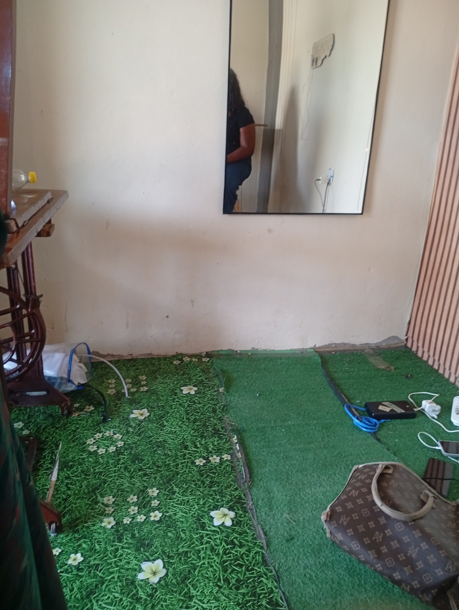 The image shows an indoor scene with a woman reflected in a mirror. The woman, wearing a dark t-shirt and blue jeans, is sitting down. The room has a light-colored wall and a floor covered with what appears to be artificial grass with floral patterns in some areas and solid green in others. To the left, there is a vintage sewing machine with a wooden surface. A white plastic bag is near the sewing machine. On the right side of the room, there is a brown designer handbag with a repeating logo pattern. Several electronic devices, including chargers and cables, are scattered on the green floor. A light pink, striped curtain is visible on the far right. The lighting suggests it might be daytime. The overall impression is of a somewhat cluttered but functional space.