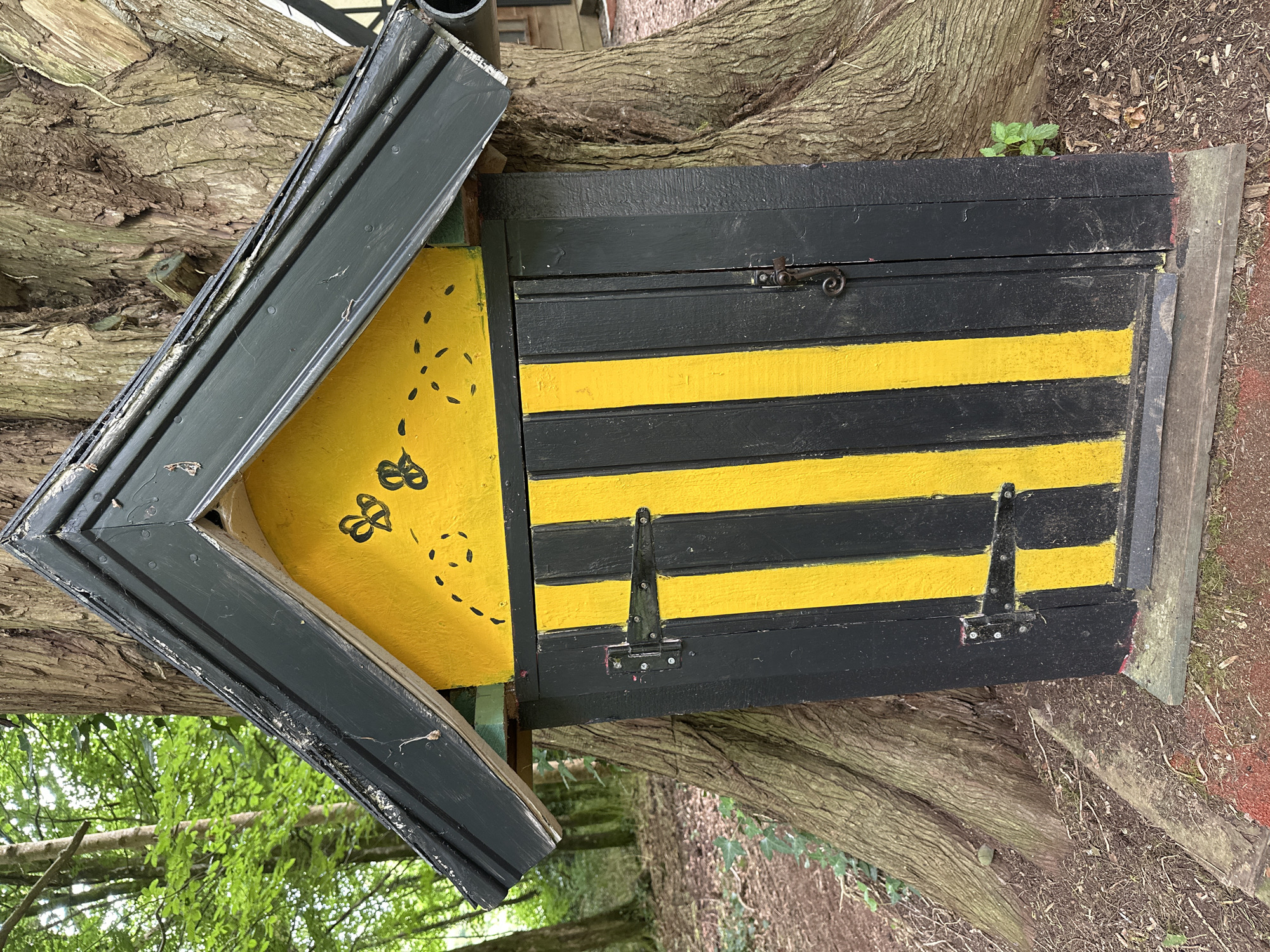 A small, dark green, gabled structure with a yellow front face, painted with black vertical stripes and two black bee silhouettes, is built against the trunk of a large tree. The structure has a dark green roof and a small, dark metal latch on its right side. The tree has rough, textured bark and is surrounded by leaf litter and sparse vegetation. The background shows a blurred image of a wooded area with green foliage and sunlight filtering through. The structure appears to be a miniature building, possibly a decorative item or a small enclosure. The scene is set in the townland of Ballycomar, County Kilkenny, Ireland.