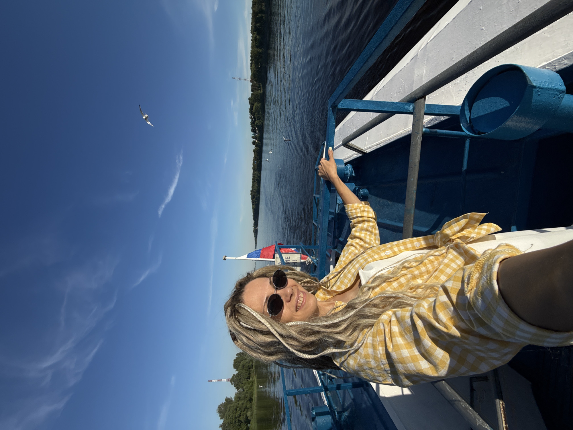 A woman is depicted on the bow of a passenger boat, navigating a wide body of water, described as being in the city of Dumino, Russia, under a clear blue sky with sparse cirrus clouds. The woman, with long blonde and white dreadlocks, is wearing dark oval sunglasses, a yellow and white checkered long-sleeve shirt over a white top, and is smiling towards the camera. Her right hand rests on the boat's blue metal railing. Behind her, a Russian flag is visible on a pole, with partially obscured Cyrillic text below it. The boat features blue railings, deck elements, and a blue cylindrical container on its white deck structure. The water is calm, exhibiting dark blue hues and surface ripples. On the distant banks, dense green treelines are visible. A prominent red and white striped lattice tower stands on the left bank, and another less distinct tower is visible on the far right bank. Several seagulls are observed flying low over the water and resting on its surface. The scene is illuminated by direct sunlight, casting shadows from the boat's structure.