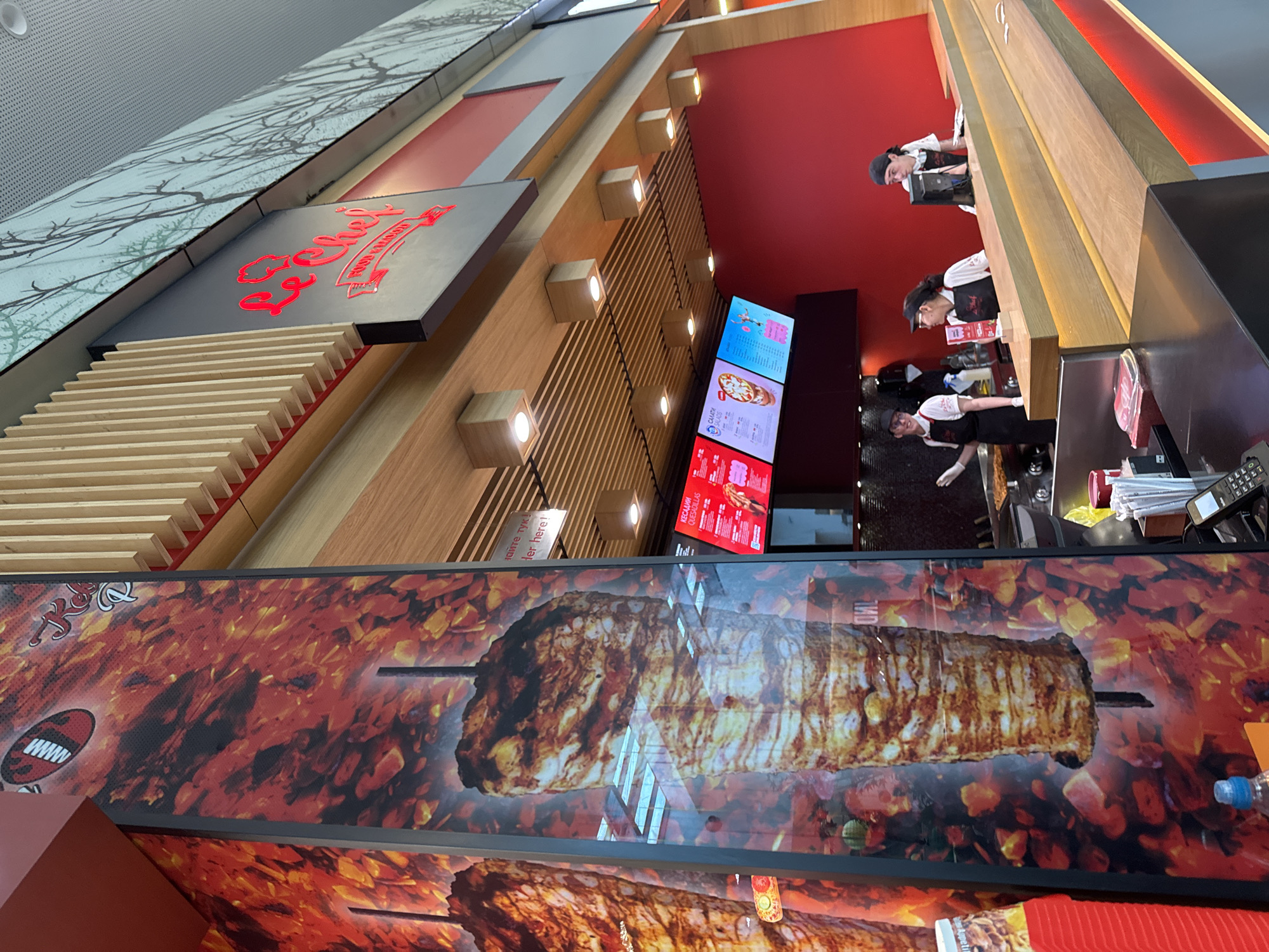 The image depicts the exterior of a "Le Chef" fast-food restaurant, likely located in Varna, Bulgaria, given the Cyrillic text visible on a menu board. The restaurant features a prominent vertical display of a döner kebab, set against a backdrop of fiery red and orange patterns. A red sign with the "Le Chef" logo, featuring a chef's hat, is visible above the counter area.
Inside the restaurant, three employees are visible behind a wooden counter. Two are wearing black aprons and white shirts, and one is wearing a black cap and glasses. The third employee, also in a black apron and white shirt, is seen from the side. They appear to be serving customers or preparing food.
Digital menu boards are displayed behind the counter, showing various food items and prices. One board shows a pizza and salad, while another displays what appears to be a dessert or drink. The interior design incorporates warm wood tones, red accent walls, and modern lighting fixtures. The overall scene suggests a busy, contemporary fast-food establishment.