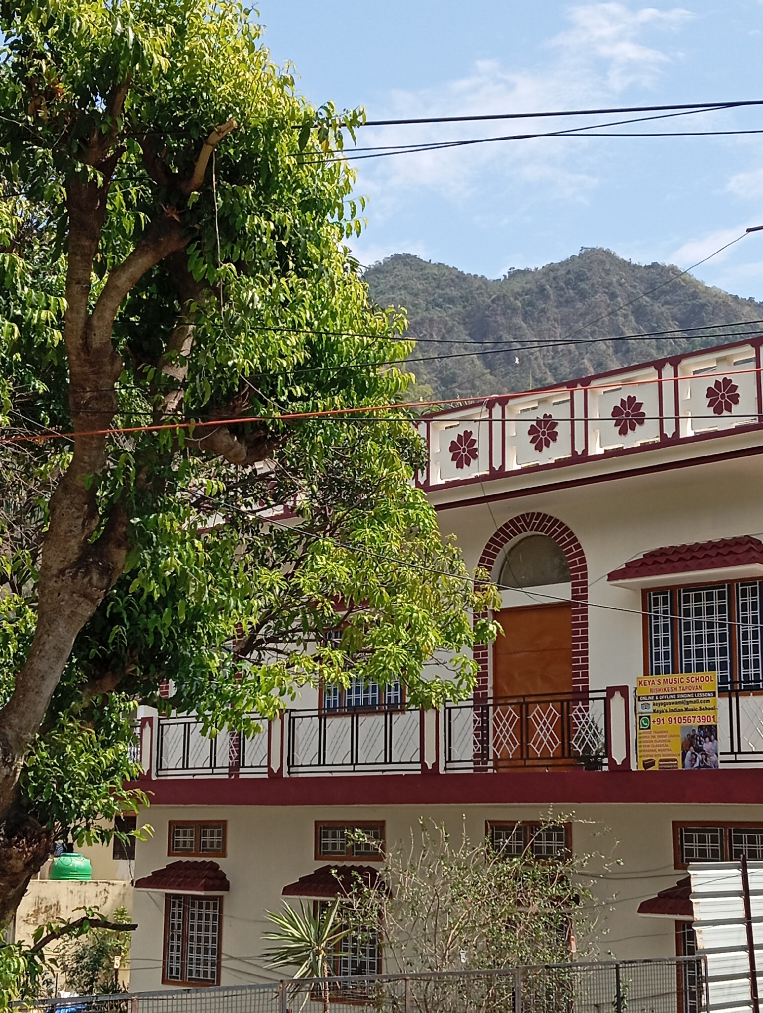 The image shows a two-story building partially obscured by a large tree with green leaves. The building is painted white with red accents around the windows, roof, and balcony. The balcony has a metal railing and a sign that reads "KEYA'S MUSIC SCHOOL RISHIKESH TAPOVAN" with contact information. Windows are visible on both floors. In the background, there are mountains. The location is likely Rishikesh Tapovan, India, based on the sign. The sky is blue with some clouds. There are utility wires visible running across the top of the image. A green water tank is visible behind the tree. A fence runs along the base of the property.