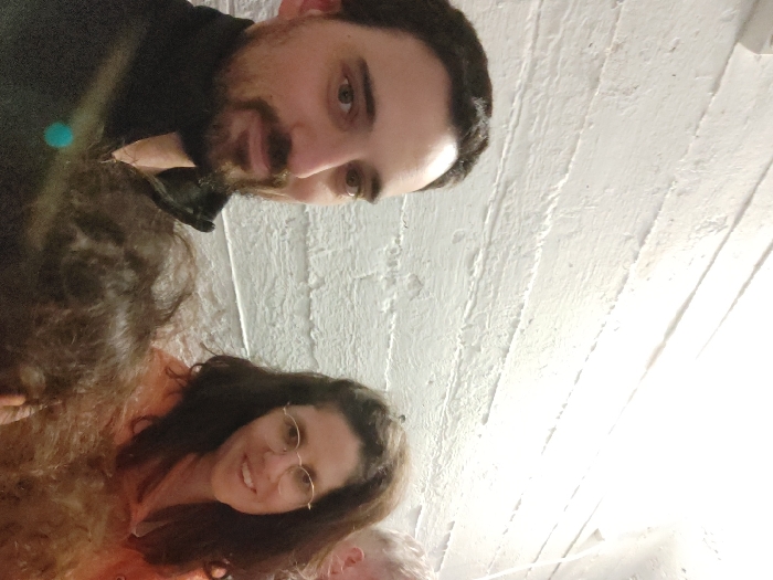 A close-up, moderately lit indoor photograph displays four individuals in an unspecified internal location, indicated as Tel-Aviv, Israel, by contextual information, though no specific visual cues confirm this. In the left foreground, a male subject with dark hair and a beard, wearing a black collared jacket, faces the camera directly. To his right, a female subject with long, dark, curly hair, wearing round-framed glasses and an orange hooded sweatshirt, smiles while looking towards the right of the frame. Between these two primary subjects, in the immediate foreground, the top of a head with dark, curly hair is partially visible. On the extreme right edge of the frame, a portion of a fourth individual's head with lighter hair is discernible behind the female. The background consists of a uniformly white, horizontally textured wall. A distinct blue lens flare is present in the lower-left section of the image. The subjects appear to be at a social gathering.