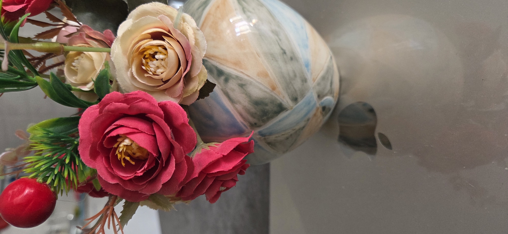 This close-up, still-life image presents an artificial floral arrangement with a decorative ceramic object in the background. The composition is filled with several faux flowers, primarily roses, and green foliage, set against a smooth, reflective surface.
The most prominent elements are artificial roses in two main color palettes. On the left and foreground, there are deep fuchsia or red roses with visible fabric petals and bright yellow-orange synthetic stamens. Alongside these are smaller, tightly-budded red roses. Interspersed are pale cream or light peach-colored roses, some with subtle pink edges, also displaying fabric textures. Green artificial leaves and spiky, needle-like green foliage are woven throughout the arrangement. On the far left, a single, bright red, smooth, spherical berry-like object adds a contrasting pop of color.
In the background, positioned to the right of the flowers, a portion of a decorative ceramic object is visible. This object has a glossy, iridescent surface with an abstract, mottled pattern in shades of pale blue, muted green, beige, and white, showing bright reflections. The overall background is a flat, light-colored, and highly reflective surface, likely a tabletop or wall, which captures soft, indistinct reflections of the ceramic piece and shows minor dust or smudges. No people, specific activities, or visible text are present. The lighting suggests an indoor setting.