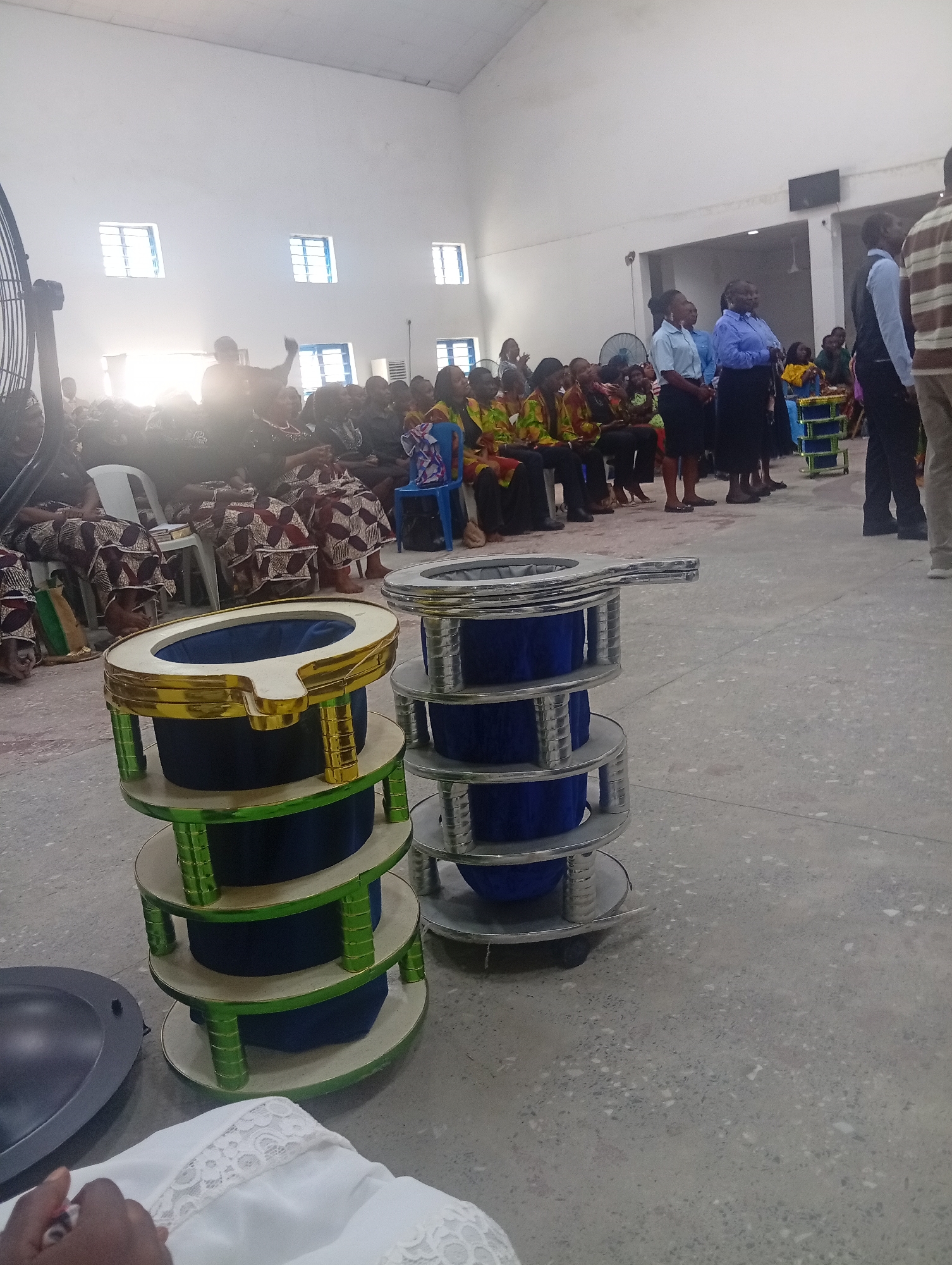 The image depicts an indoor gathering, likely a church service or religious event, held in Kason, Nigeria. The room has high, white-painted walls and a concrete floor. Natural light streams in from several windows on the left side of the room. In the foreground, two tiered, cylindrical objects with blue fabric lining and metallic rims are prominently displayed. The one on the left has a green and gold decorative trim. A dark, circular object, possibly a bowl or platter, is located to the left of these tiered items. A person's arm, adorned with white lace fabric, is visible at the bottom of the frame, suggesting they are seated.
Further back, a congregation is seated on chairs. Many individuals are wearing brightly colored, patterned traditional African attire, primarily women. Several individuals in the middle ground are standing, some appearing to be involved in leading or organizing the event. These individuals are dressed in more formal attire, including shirts and skirts/trousers. A large industrial fan is situated in the far left background. The overall atmosphere suggests a community event or service taking place in a modest, communal space.
