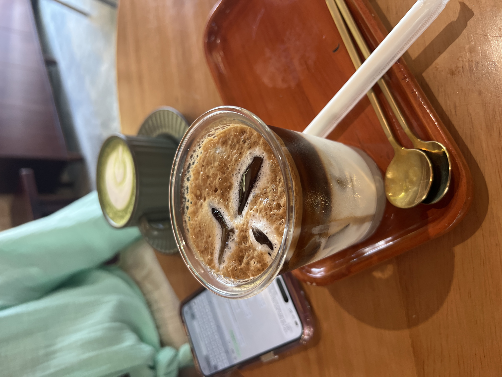 The image shows a table setting with a brown wooden table and a light green cushioned seat visible in the lower left. On the table, there's a cup of iced coffee with brown ice cubes and a foamy top, positioned next to a smartphone with a screen displaying a chat interface. To the left of the coffee, there's a green ceramic cup with a green liquid, possibly matcha tea, placed on a saucer. A brown wooden tray holds two golden spoons and a white straw, likely intended for the drinks. The lighting suggests an indoor setting, and the overall composition indicates a casual dining or cafe scene. The location is Nha Trang Ward, Vietnam.