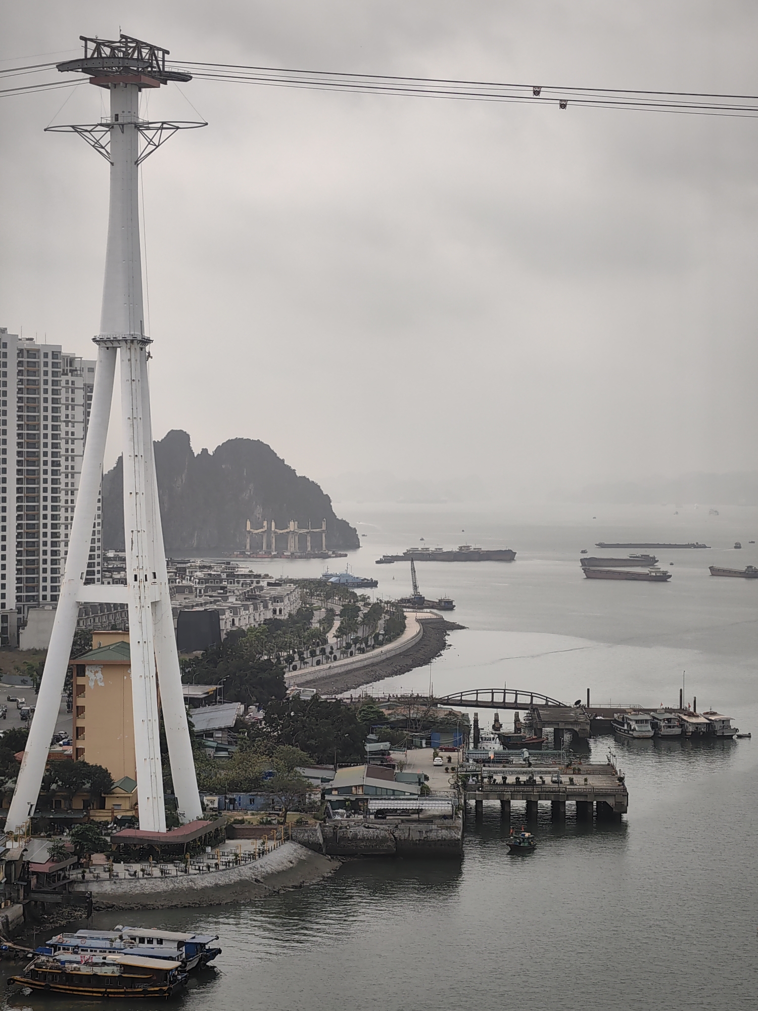 The scene captures an elevated view of Bai Chay Ward, Vietnam, during an overcast day. A prominent white cable car tower dominates the left foreground, with cables extending across the frame. Adjacent to the tower are high-rise buildings, followed by a coastal road lined with trees and buildings. Beyond the coast, a bay is populated with numerous cargo ships and boats, some anchored near an island. A pier with several docked boats is situated in the lower right. A small boat is moving on the water in front of the pier. There is an open-air restaurant with tables and chairs visible.