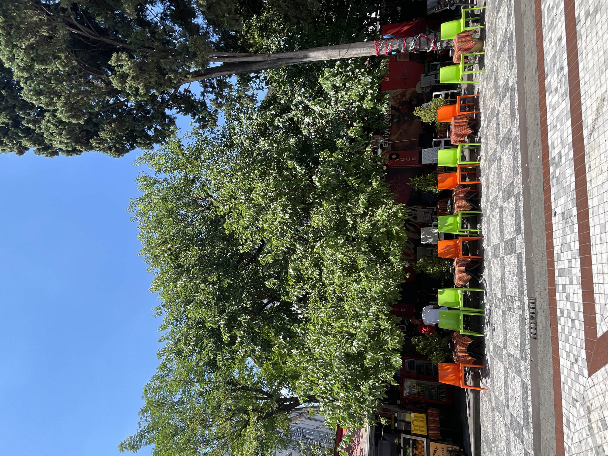 The image depicts an outdoor seating area in front of a cafe or restaurant on a bright, sunny day. The scene is set in the city of Kadıköy, Turkey, likely on a pedestrian walkway. In the foreground, a patterned tiled sidewalk leads towards the seating area. Directly behind the sidewalk, a row of small, round tables are set up, each covered with a colorful, patterned tablecloth. Arranged around these tables are an alternating pattern of bright orange and lime green plastic chairs.
Behind the tables and chairs, a building with a red awning is visible. Various items are displayed on shelves and in refrigerated units in front of the building, suggesting it is a food or drink establishment. A man in a white shirt is standing behind a counter inside the establishment. Lush green trees, one large and dense with foliage and another taller, more slender tree with needle-like leaves, provide ample shade over the seating area and extend towards the bright blue sky. The overall impression is a vibrant and inviting outdoor space, typical of a European-style cafe in a bustling urban environment.