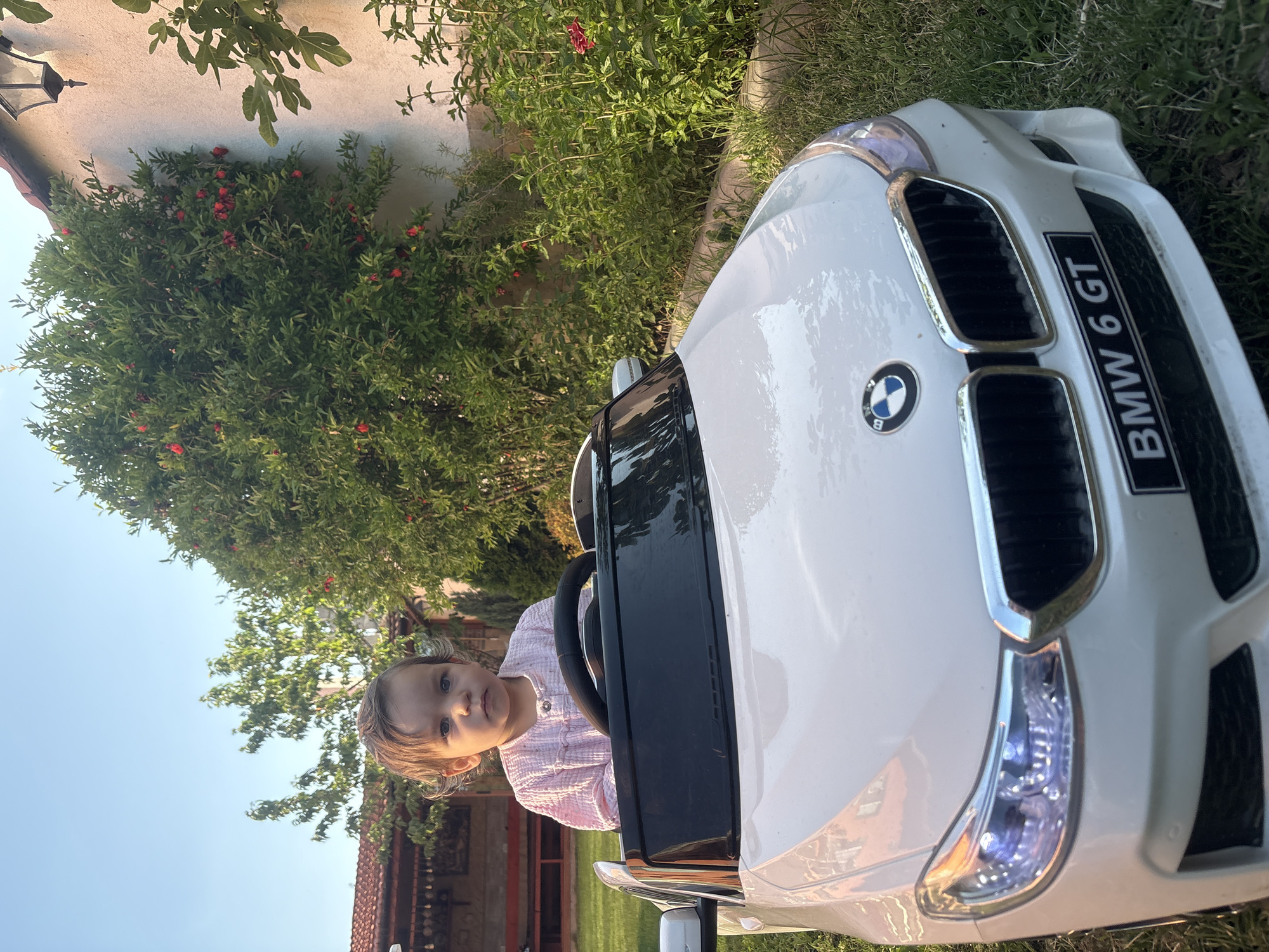 A young child sits in the driver's seat of a white toy BMW 6 GT. The child, wearing a pink shirt, looks at the camera with a curious expression. The toy car is parked on grass, with a lush green bush and a building with a red-tiled roof visible in the background. A clear blue sky suggests it is daytime. The toy car has the "BMW 6 GT" text displayed on the front grille. The overall scene is set outdoors, likely in a garden or yard, with elements of nature and a residential structure.