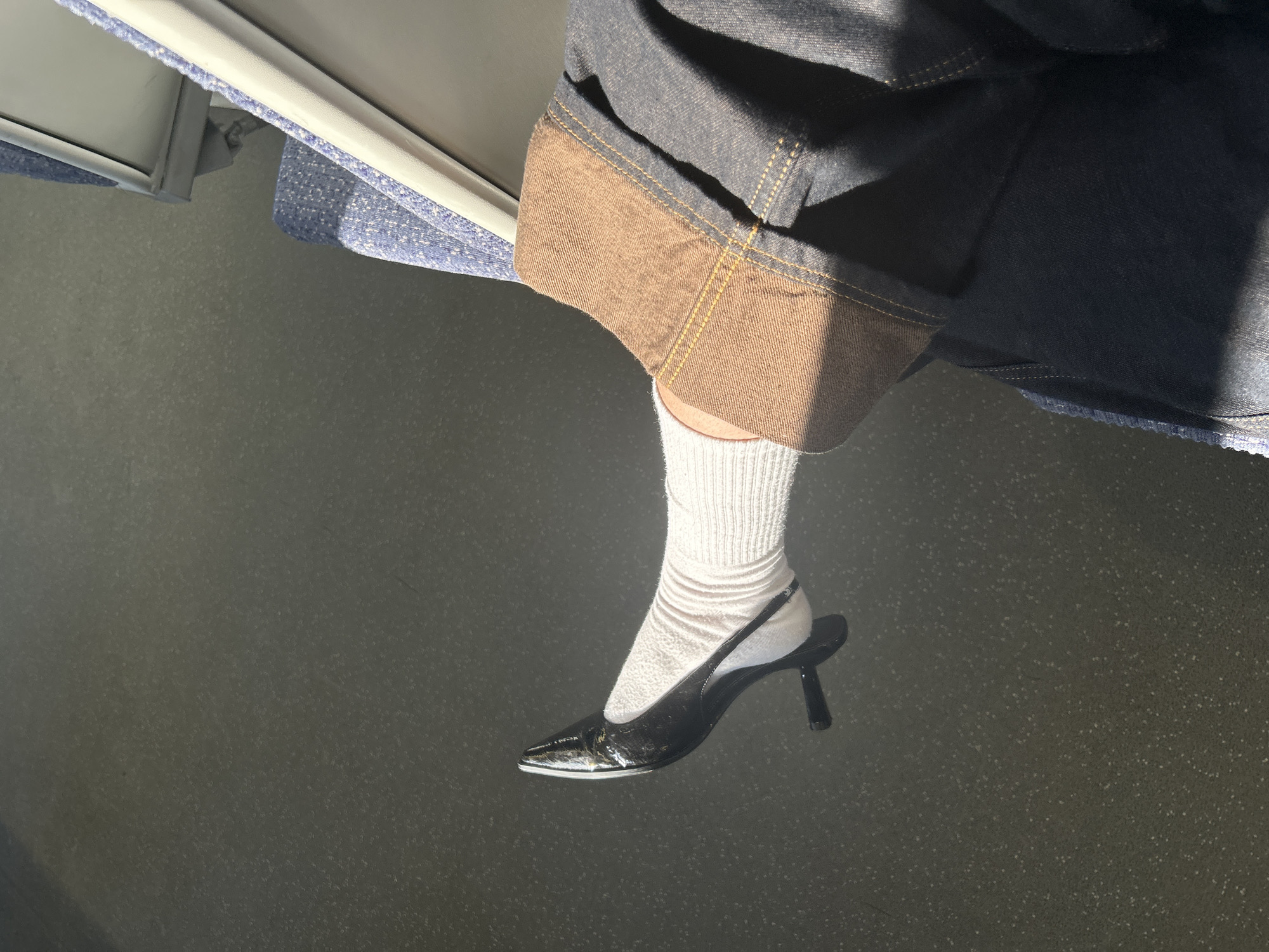 The image depicts a person seated on what appears to be a train or bus, judging by the blue seat fabric and the window frame visible in the upper left corner. The person is wearing denim shorts with a brown, cuffed hem. They are also wearing a white, ribbed sock and a black patent leather slingback heel. The floor is gray with a speckled texture. The lighting suggests it is daytime, possibly inside a vehicle. There are no identifiable features to pinpoint the exact location in Hertsmere, United Kingdom.