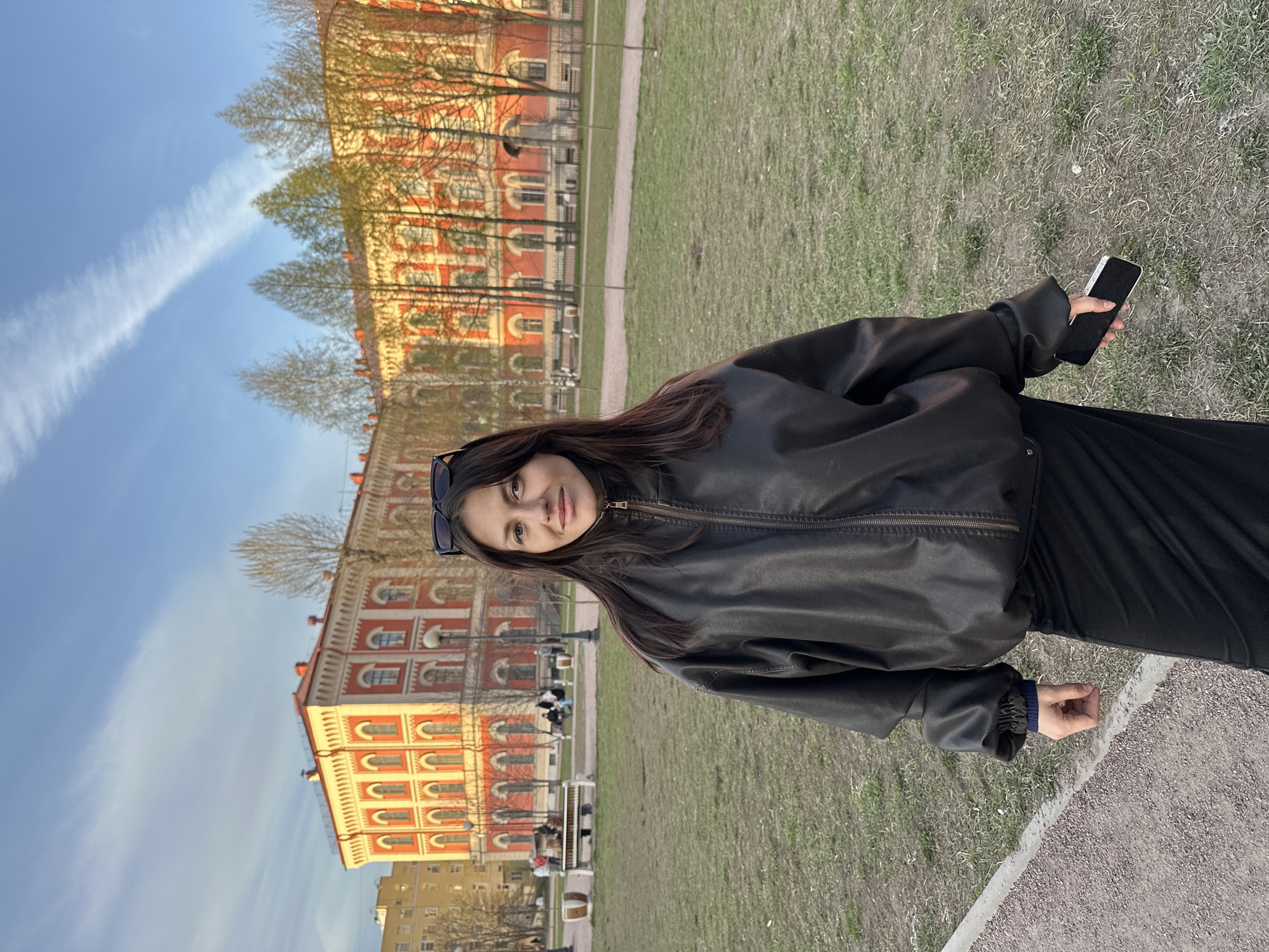 A young woman stands outdoors in what appears to be a park or green space in Saint Petersburg, Russia, posing for a photograph. The image is taken from a slightly low angle, showing her from the waist up, and she is looking directly at the camera with a neutral to slight smile. She has long, dark hair and is wearing a black leather jacket over a dark top or dress. Sunglasses are pushed up onto her head. In her right hand, she holds a white-framed smartphone, while her left hand is loosely clenched by her side.
Behind her, the scene features a grassy area with a dirt or gravel path. In the mid-ground, there are several park benches, with a few indistinct figures seated on them. The background is dominated by large, ornate buildings with red and cream-colored facades, featuring numerous windows and classical architectural details, consistent with the style found in Saint Petersburg. Some bare trees and trees with sparse, light green foliage are scattered throughout the park, suggesting early spring.
The sky is a clear, bright blue, with a prominent white contrail stretching diagonally across it. The lighting suggests late afternoon or early evening, with a warm, golden light illuminating the buildings. No visible text is present in the image.