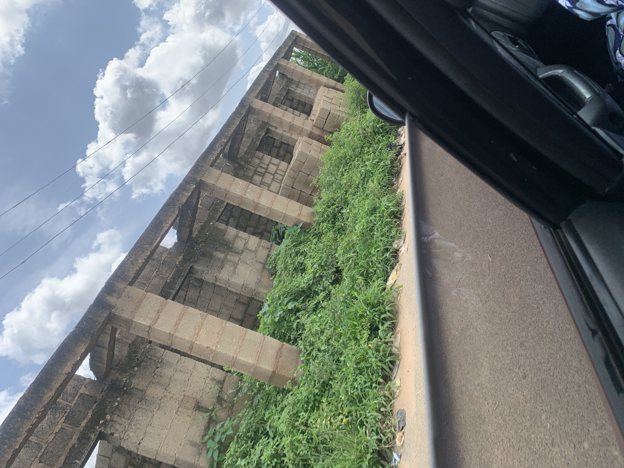 The image shows a partially constructed building made of concrete blocks and beams, with some of the blocks stacked on the ground inside. The structure is overgrown with lush green vegetation, which also covers the ground around it. The scene is viewed from the interior of a vehicle, with the edge of the car door and window visible in the foreground. A road is visible in front of the building, and the sky above is partly cloudy with clear blue patches and thin white clouds. There are utility wires running across the sky. The overall impression is of an abandoned or unfinished construction site reclaimed by nature. The location is identified as Unknown, Unknown.