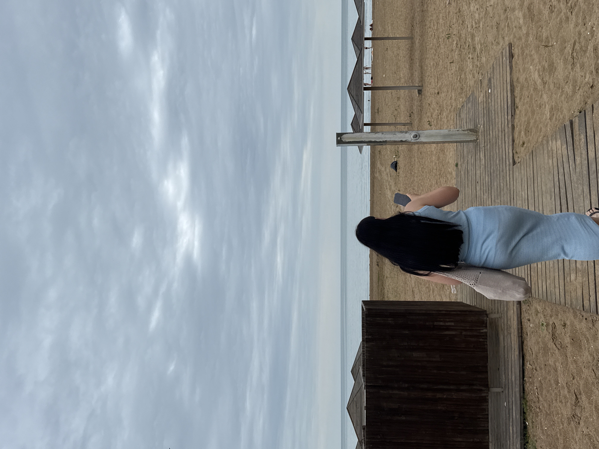 A woman with long black hair, wearing a light blue dress and carrying a beige tote bag, walks on a wooden boardwalk towards the sea on an overcast day at a beach in Derbent, Russia. She is holding a black smartphone in her right hand. In the background, there are several wooden beach umbrellas and a few people scattered on the sandy beach. The calm sea stretches to the horizon under a cloudy sky.
