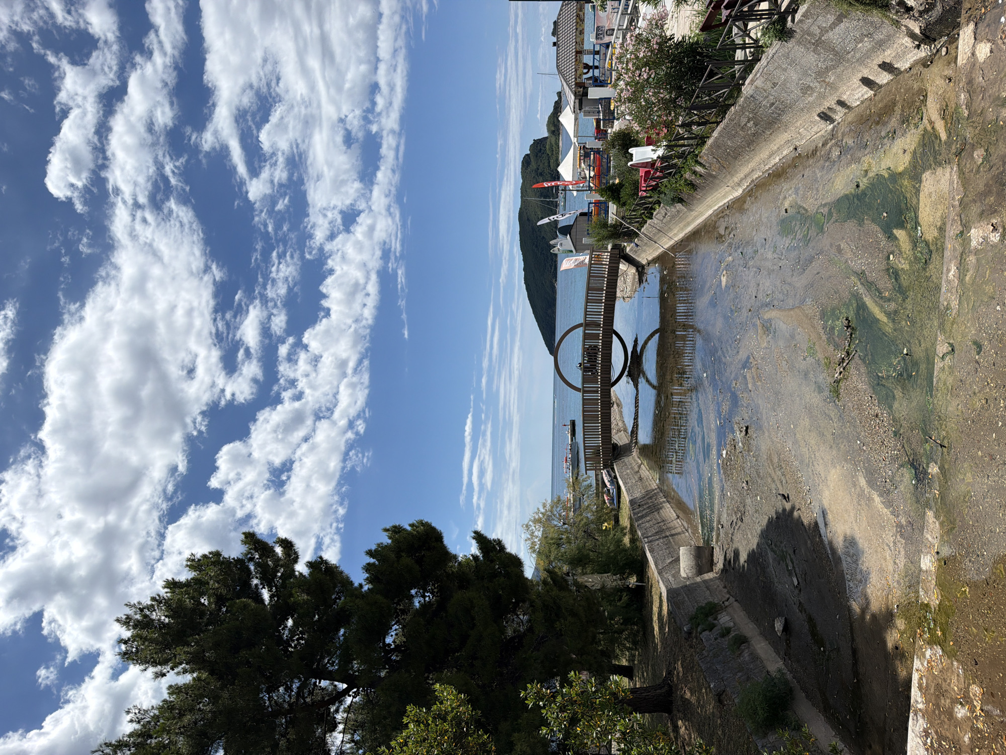 A wooden bridge with a circular feature spans across a dried-up canal. The canal has concrete banks and a muddy, uneven bottom with patches of green algae. On the left, a large evergreen tree with dark needles is prominent. In the background, the Adriatic Sea is visible, with a green, hilly island in the distance. Several boats, including sailboats and smaller vessels, are docked or anchored near the shore. Along the right bank of the canal, there are various structures, including tents, a small building with a tiled roof, and some colorful slides. A black bird is standing in the canal. The sky is blue with scattered white clouds. The scene appears to be in Budva, Montenegro, given the distinctive island and coastal features often seen in the area.