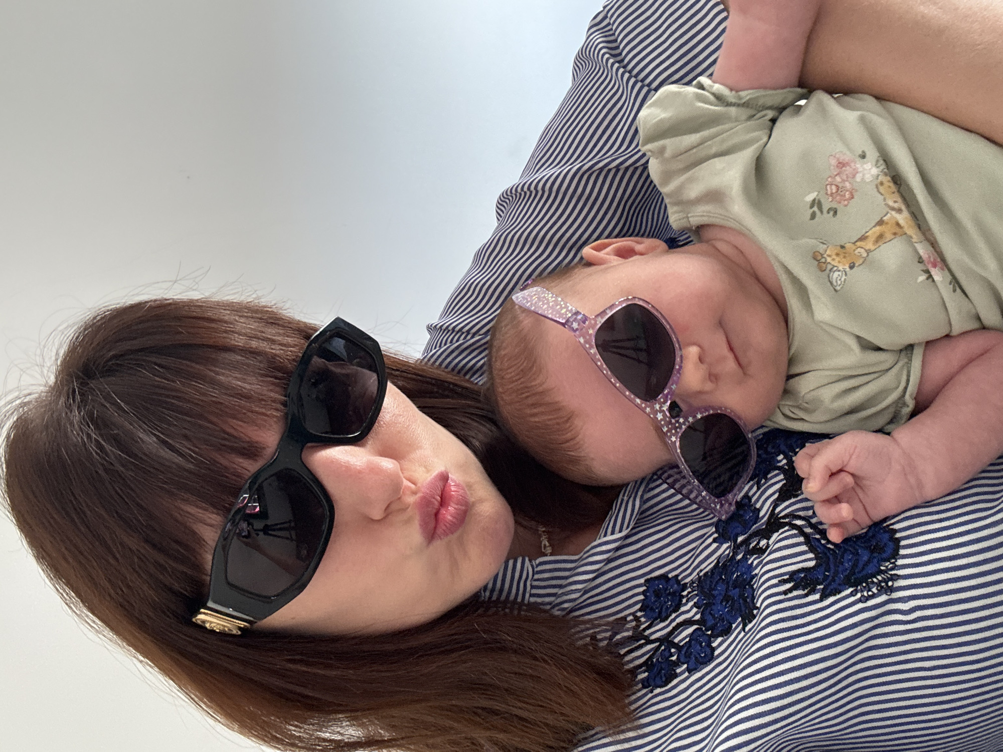 A woman and a baby, both wearing sunglasses, are close together, looking into the camera. The woman has her lips pursed, and her face is partially obscured by large black sunglasses. She has long brown hair. The baby is wearing small, sparkly purple sunglasses and is nestled in the woman's arms. The baby is wearing a light green outfit with a printed giraffe and flowers. Both are wearing striped shirts, the woman's is dark blue and white, and the baby's is also dark blue and white. The background is a plain white wall. The image appears to be taken indoors. The overall mood is playful and lighthearted. There is no visible text. The location context provided is Isola delle Femmine, Italy, which provides a possible geographical setting but no direct visual cues in the image support or contradict this.