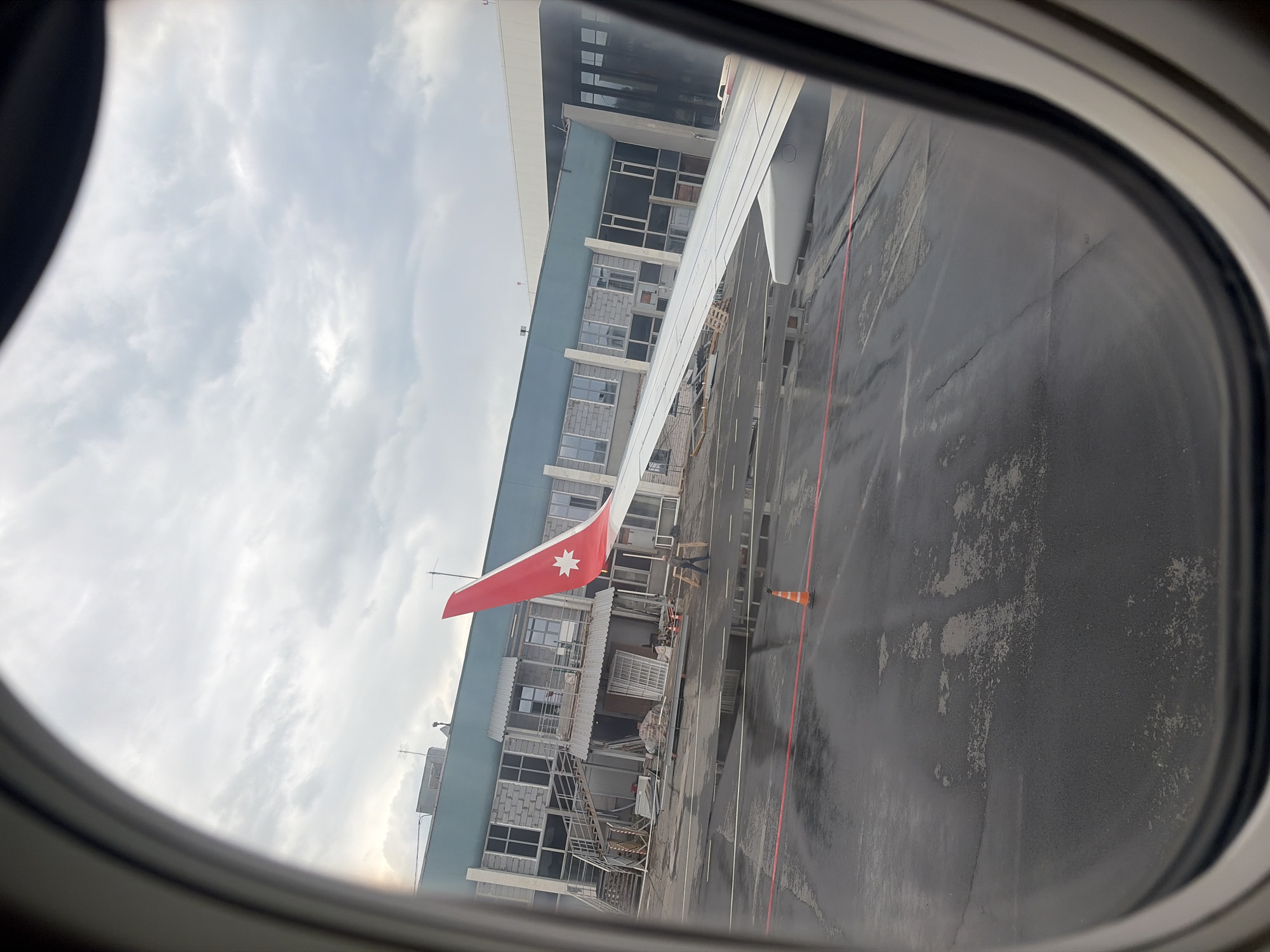 The image, taken from an airplane window, shows a parked aircraft at an airport terminal under an overcast sky. The photo is oriented vertically, rotated 90 degrees counter-clockwise.
The foreground is framed by the dark, rounded outline of the aircraft window. Through it, the white wing of the airplane extends prominently across the left side of the frame. The wing's tip is painted a distinctive red, featuring a clear white five-pointed star, which is characteristic of Georgian Airways livery.
Adjacent to the aircraft wing, an airport terminal building is visible, spanning horizontally across the middle of the image. The building's facade consists of multiple large windows, some with reflections, and sections of exposed grey concrete blocks, suggesting an older structure or parts undergoing renovation. A light blue band runs along the upper portion of the building. Antennas are perched on the roof.
The tarmac below appears dark and somewhat wet in areas, hinting at recent rainfall or damp conditions. Orange traffic cones are placed on the tarmac, and red painted lines indicate operational boundaries or pathways. A single individual, dressed in dark clothing, can be faintly seen standing near the base of the wing and the terminal building.
The sky is uniformly cloudy and grey, diffusing the daylight. The overall lighting suggests it is daytime, possibly early morning or late afternoon due to the subdued light. There is no discernible text visible on the aircraft or the building. Given the context of Новое Мартьяново, Russia, the image depicts a Georgian Airways aircraft at a Russian airport.