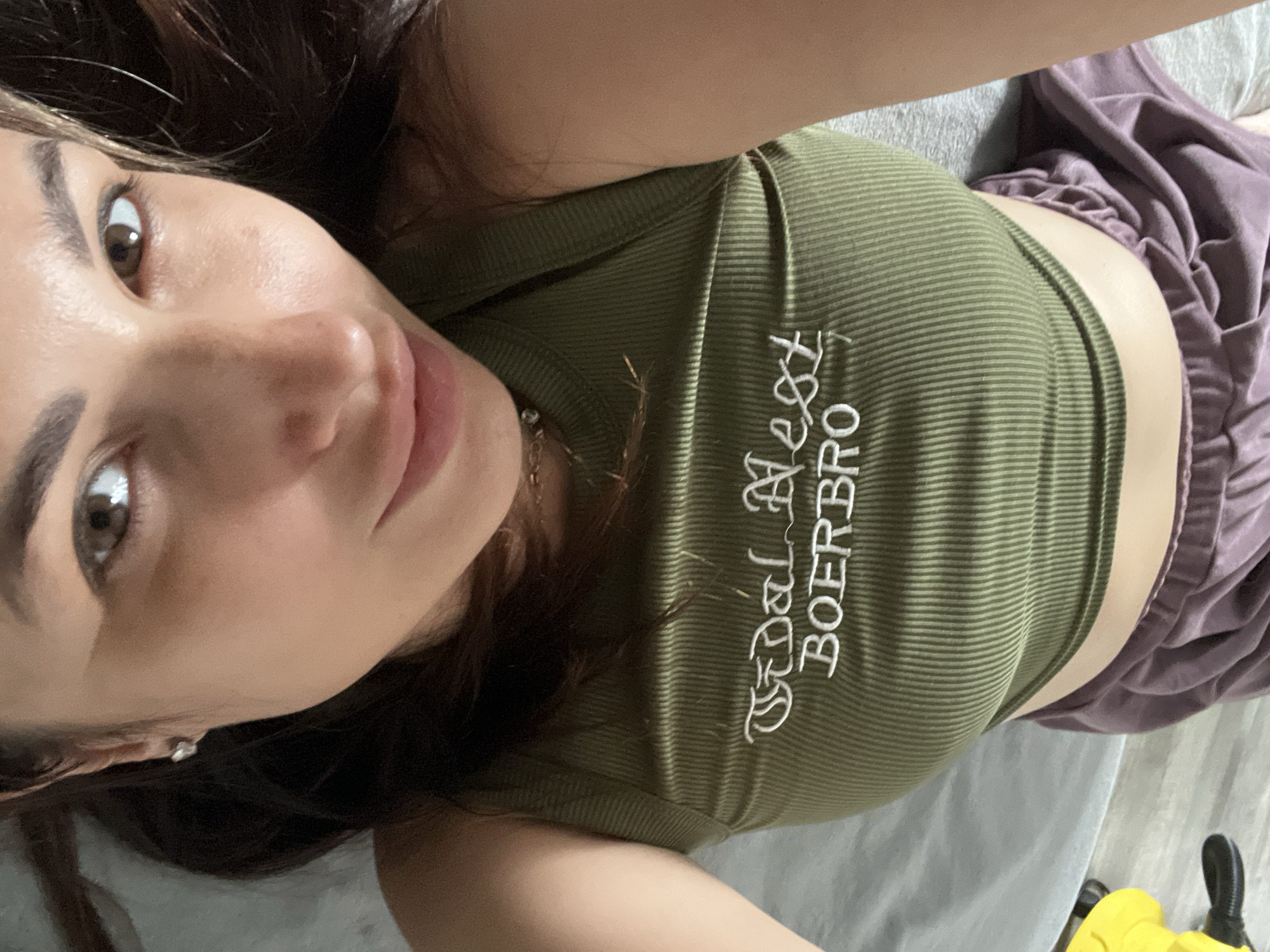 A person is lying on their back on a gray surface, wearing a green ribbed tank top and purple shorts. White text on the tank top reads "BOERBRO GOAL BEST". The person's face is turned towards the camera, with their eyes open and looking upwards. Their hair is dark brown and extends to the sides. A yellow object is visible in the upper right corner of the image. The scene appears to be indoors, possibly a bedroom or living space.