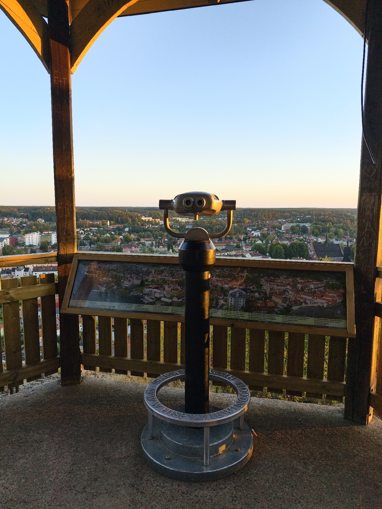 An eye-level, medium shot captures a public viewing station within an open-air wooden structure. The station features a coin-operated, mounted binocular viewer, a large information panel displaying a map of the town with numbered points of interest, and a decorative metal base with inscriptions. The binocular viewer is positioned centrally, facing the town below. The information panel, situated behind the viewer, showcases a detailed map of Söderköping, Sweden, with Swedish text identifying landmarks. A wooden railing encloses the viewing platform, offering a clear vista of the town. The town of Söderköping, characterized by its red-tiled roofs and dense vegetation, stretches out to the horizon under a clear, pale blue sky. The warm, golden light of a setting sun casts long shadows and illuminates the wooden structure and parts of the town. The foreground consists of a textured concrete or stone floor. The scene is devoid of any human presence.