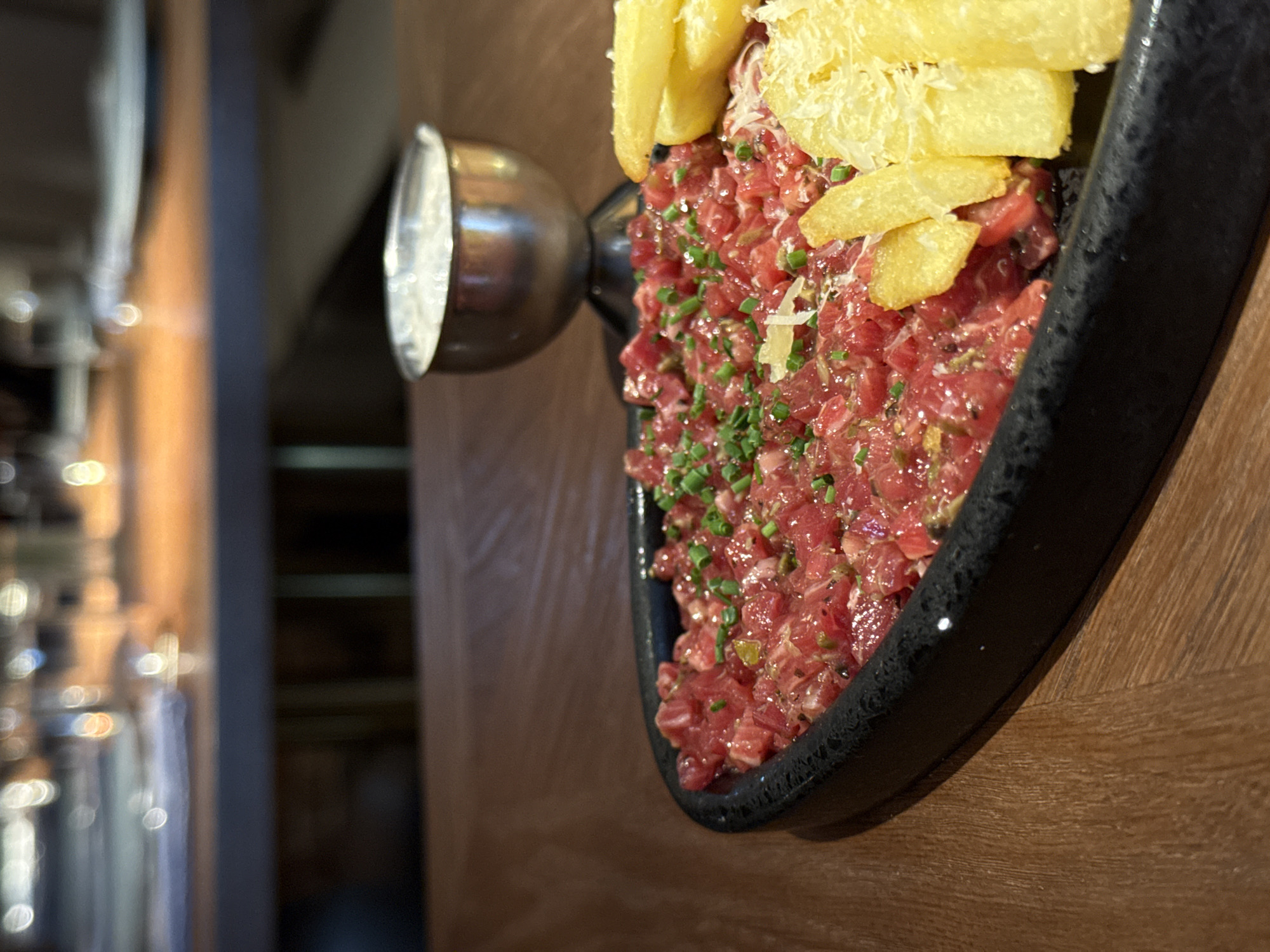 A close-up, eye-level shot focuses on a dish of raw chopped meat, likely steak tartare, presented in a black, textured bowl. The meat is finely diced and mixed with what appears to be finely chopped chives and possibly capers, giving it a speckled green and white appearance against the deep red of the meat. A few pieces of golden-brown, thick-cut fries are perched on top of the tartare, along with a sprinkle of grated cheese. To the left of the dish, a metallic object with a reflective, polished surface is visible. Its curved shape and bright reflection suggest it might be part of a light fixture or decorative element. The background is softly blurred, showing a wooden surface on the right and a darker, out-of-focus area with vertical lines on the left, possibly hinting at shelves or architectural features within an establishment. The lighting appears warm and artificial, suggesting an indoor setting, likely a restaurant or bar, and potentially evening time due to the soft glow. There is no visible text.