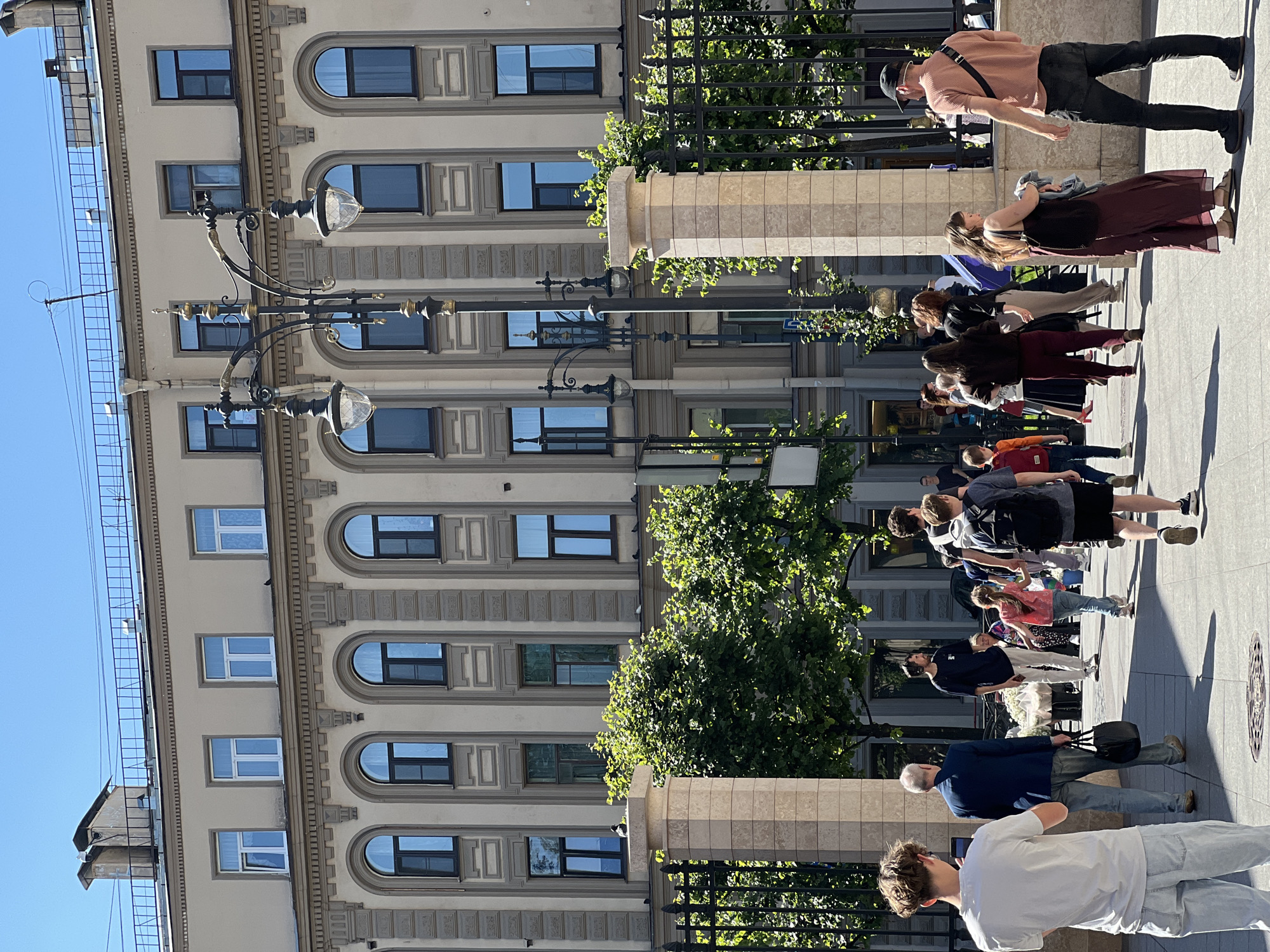 The image depicts a street scene in Saint Petersburg, Russia, with a grand building in the background characterized by multiple arched windows and ornate architectural details. A group of people are walking on a sidewalk in the foreground, some are engaged in conversation, others are looking at their phones, and a few are walking together. The weather appears to be sunny with a clear blue sky. The scene is framed by a fence and stone pillars on the right side, and lush green trees are visible behind the fence. A vintage street lamp stands prominently in the mid-ground, adding to the historical ambiance of the city. The overall atmosphere is lively and suggests a typical urban environment on a bright day.