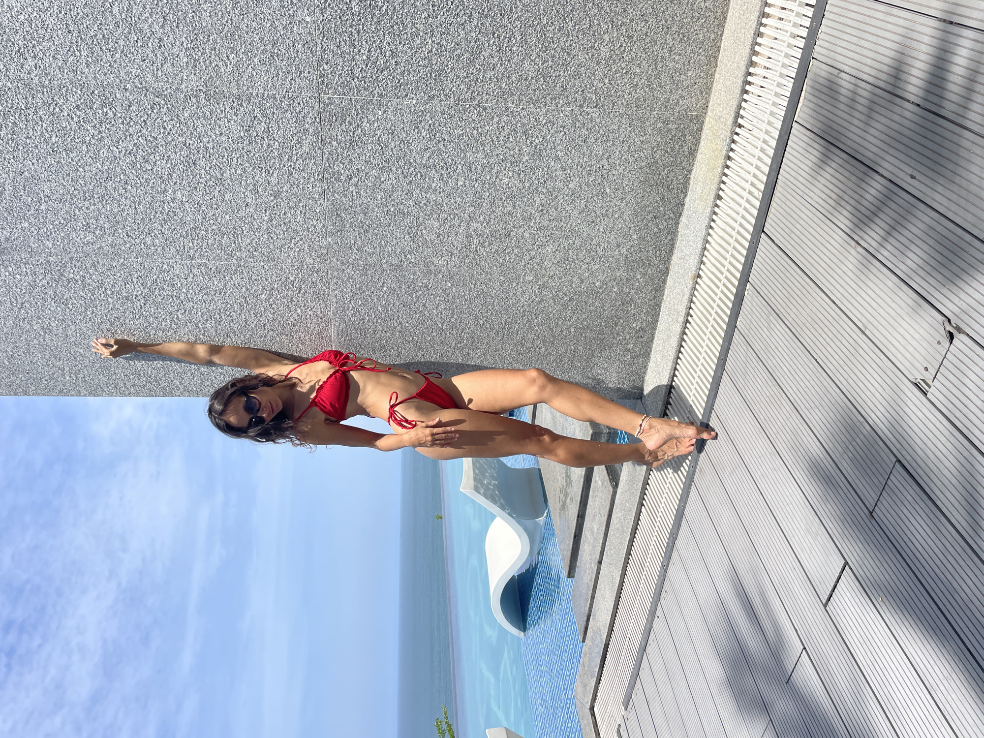 A woman in a red bikini stands leaning against a textured gray wall next to a swimming pool. She is looking down and has one arm raised, touching the wall. The pool appears to be an infinity pool overlooking the ocean. In the foreground, there is a wooden deck. The sky is blue with some scattered clouds. The image appears to have been taken in Mui Ne, Vietnam, a coastal city known for its beaches and resorts.