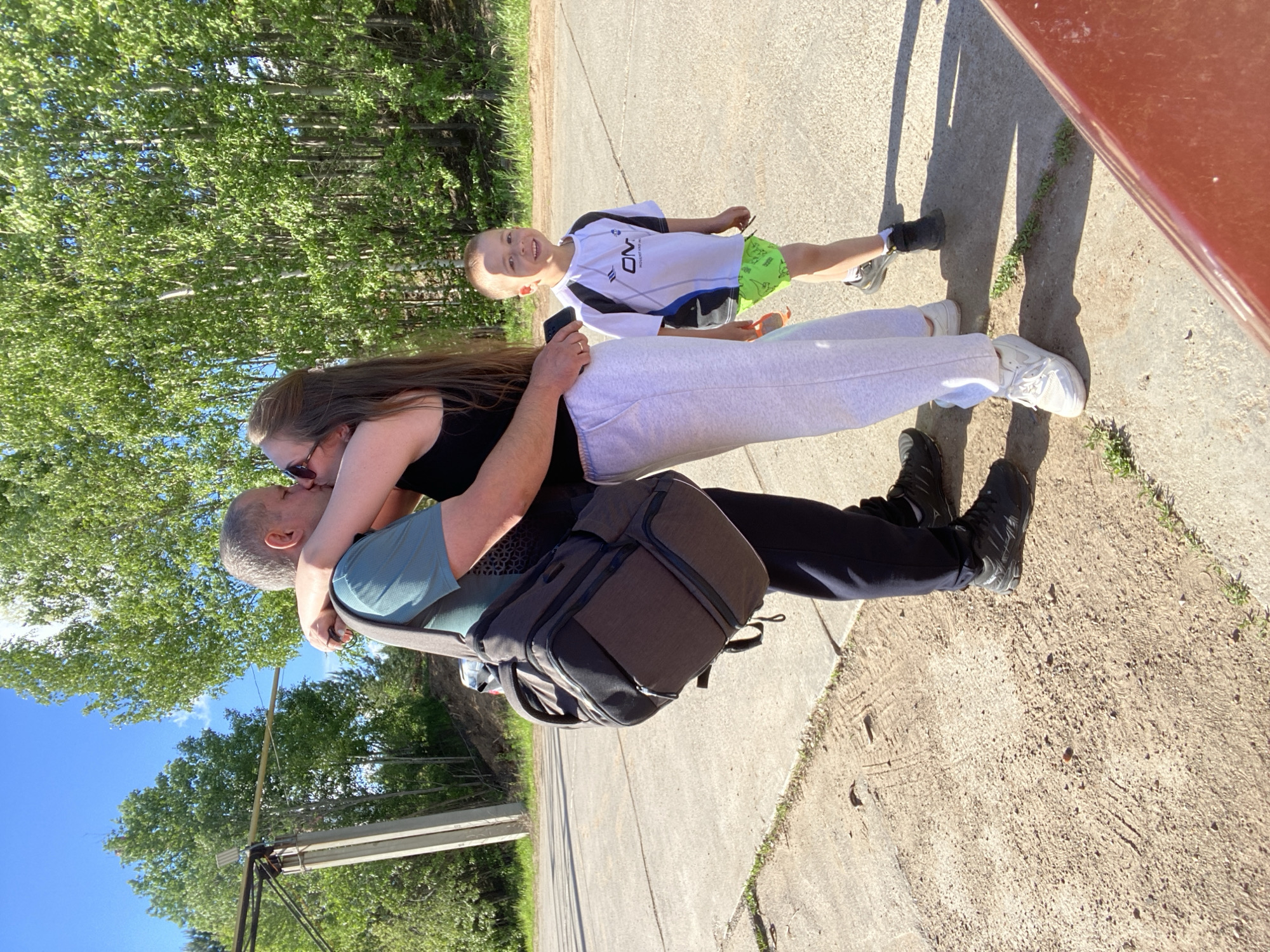 A man, woman, and child are outdoors on a paved surface near trees. The man is wearing a blue-green shirt and a backpack. The woman has long brown hair, is wearing sunglasses and a black tank top and grey sweatpants. They are embracing. The child, wearing a white shirt with a design and green shorts, is standing slightly apart and to the side. The location is possibly in the city of Chaikovsky, Russia.