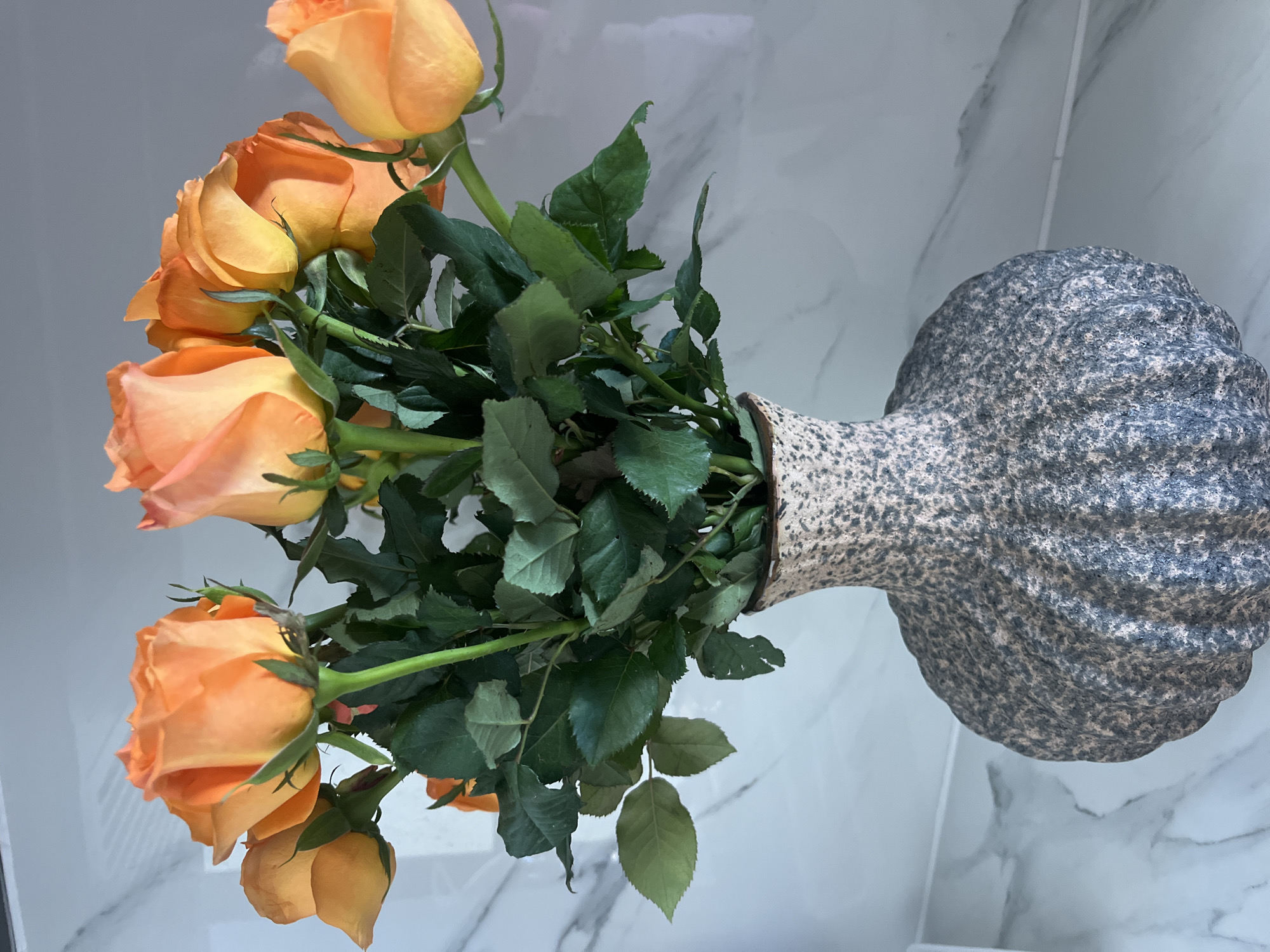 A vase of orange roses is displayed against a marble-patterned background. The vase is textured with a speckled pattern of gray and beige. The roses are a vibrant orange hue with green stems and leaves. The lighting is soft, casting subtle shadows. The scene appears to be indoors, possibly in a home or establishment in Bac Nha Trang Ward, Vietnam, given the specific location detail requested. No people are present in the image.