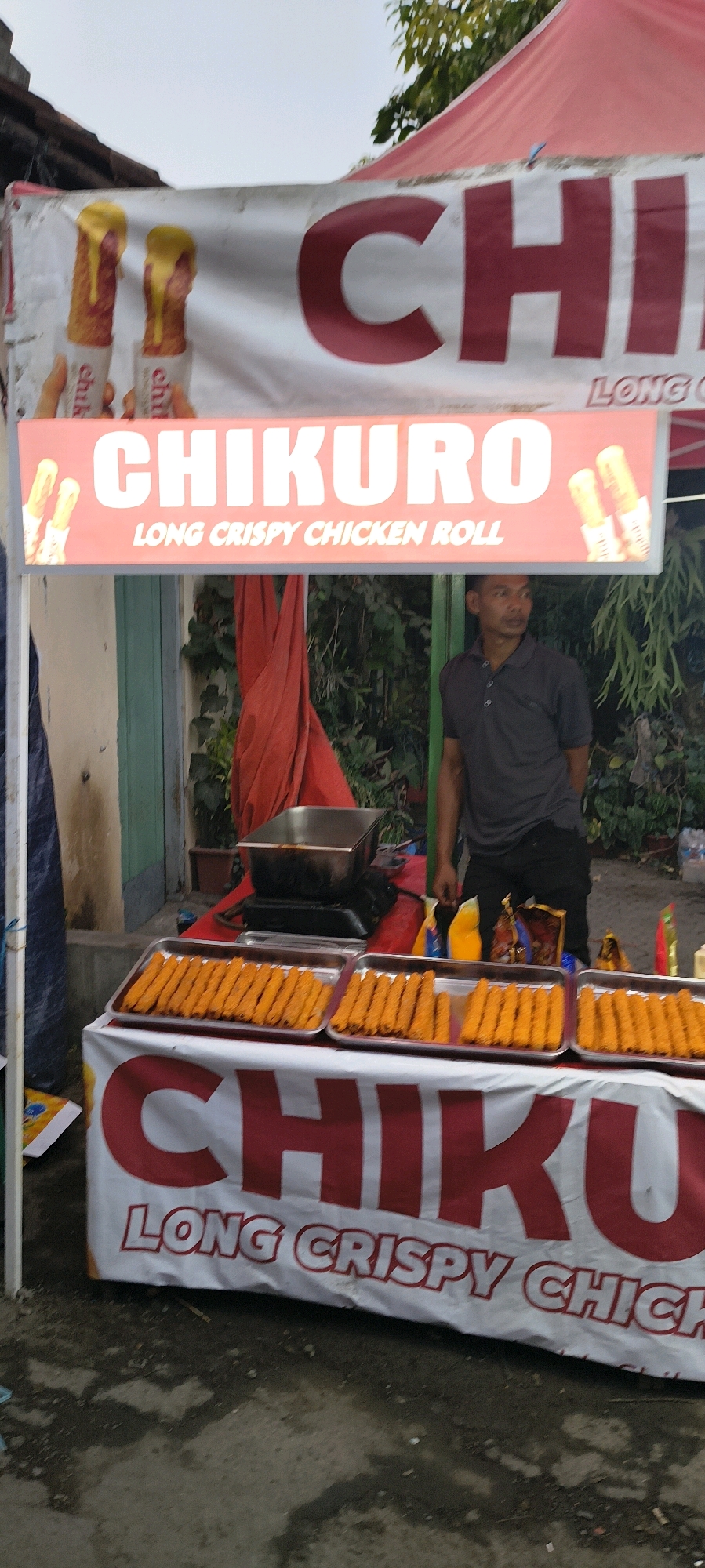 An outdoor food stall displays "CHIKURO LONG CRISPY CHICKEN ROLL" on a red and white banner. Two trays of fried chicken rolls are arranged on a table covered with a white tablecloth featuring the same logo. A man in a dark grey polo shirt and dark pants stands behind the table, looking to his right. Behind him, a red tarp is visible, along with greenery and a building. The ground is a rough, paved surface. The stall appears to be located in Trusmi Wetan, Indonesia, given the context provided.
