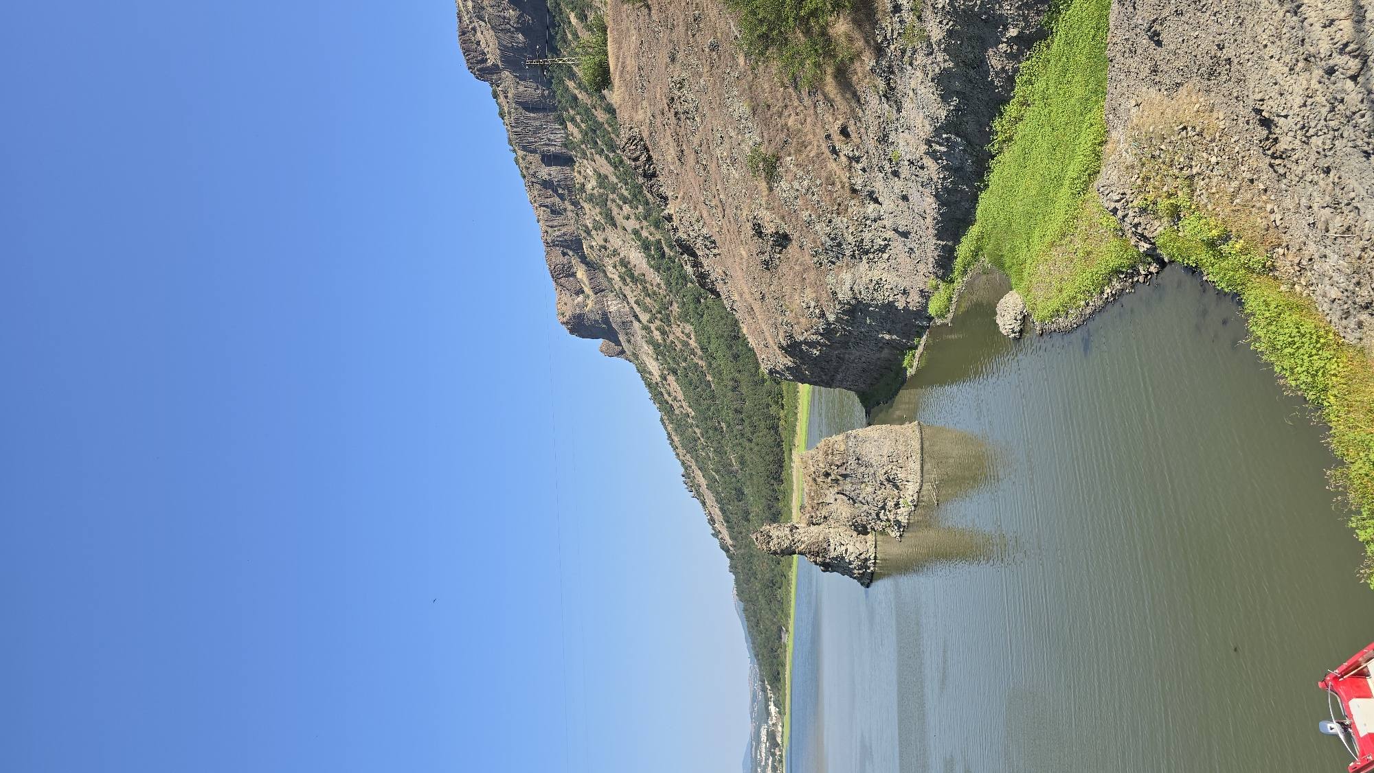 The image depicts a serene natural landscape featuring a body of water with rocky formations. In the foreground, the calm water reflects the clear blue sky. Two prominent rock formations rise from the water, one appearing as a solitary, weathered pillar and another more substantial, craggy island. The shoreline on the right is characterized by steep, rocky slopes covered in dry vegetation and interspersed with vibrant green foliage. Further in the distance, a verdant hillside rises, densely populated with trees. The uppermost portion of the image is dominated by a vast expanse of clear blue sky. The location is identified as the Lisitsite area of Bulgaria. There are no people or animals visible in the image, nor any discernible actions or interactions. The scene is one of natural geological formations and a tranquil aquatic environment under a bright sky.