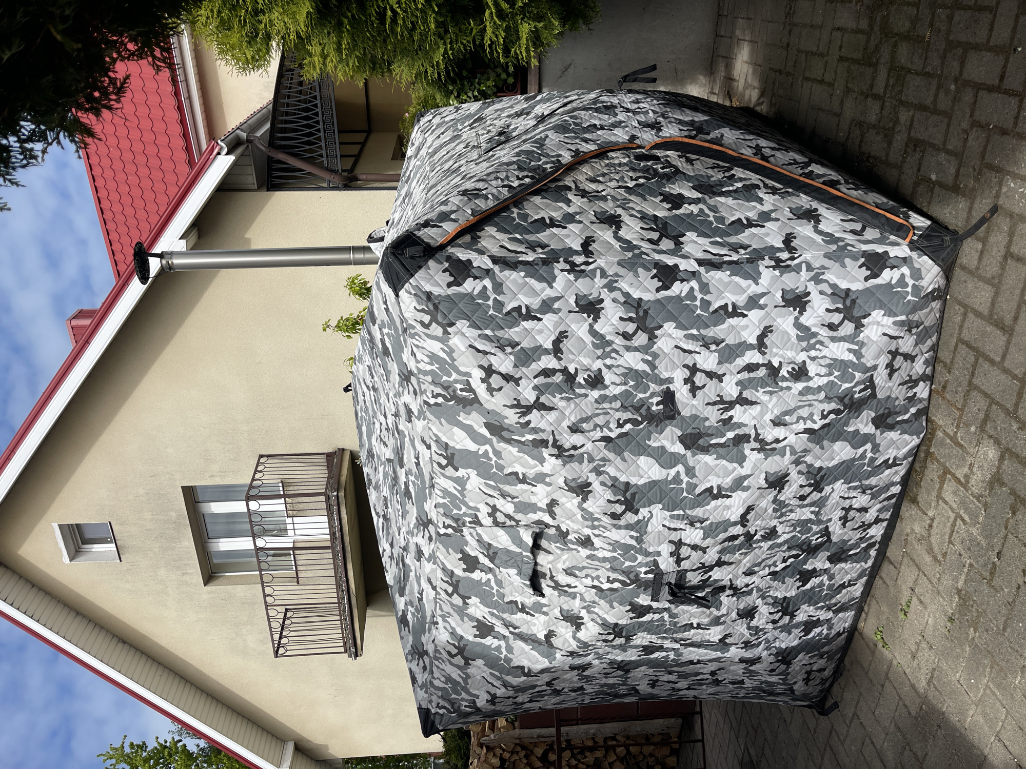 The image depicts a camouflage-patterned tent pitched on a paved surface, likely a patio or driveway, adjacent to a two-story building. The building has a red tiled roof, beige walls, and a balcony with a decorative wrought-iron railing. A metal chimney pipe extends from the building's wall near the roofline. A pile of firewood is visible near the base of the tent. Vegetation is present around the building, including a shrub or tree partially visible in the background. The sky is visible in the upper left corner, with blue sky and some clouds. The image is taken outdoors, likely in daylight. The specific city and region are unknown.