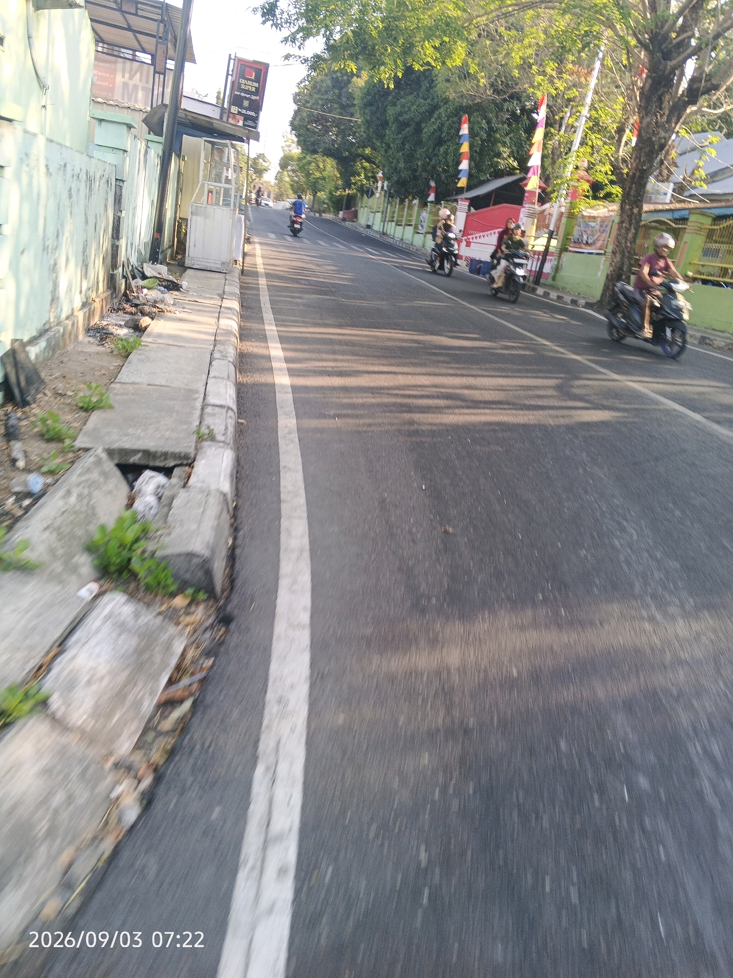 The image captures a street scene in Pare-Pare, Indonesia, on September 3, 2026, at 07:22. The perspective is from a moving vehicle, likely a motorcycle, looking forward and slightly to the right. The road is paved, with a white line marking the lane divider. On the left side of the road, there is a weathered, light green wall, a concrete sidewalk with some broken sections, and a drainage ditch containing debris and vegetation. A small, white kiosk-like structure is visible near the wall. Further down the road, on the right, trees line the street, casting shadows on the pavement. Several people on motorcycles are present. One person on a motorcycle, wearing a maroon shirt and a helmet, is riding in the right lane, moving away from the viewer. In the middle distance, three other individuals on motorcycles are traveling in the same direction. In the background, buildings and flags are visible, suggesting a populated urban or suburban area. The lighting indicates it is early morning or late afternoon, with sunlight filtering through the trees.
