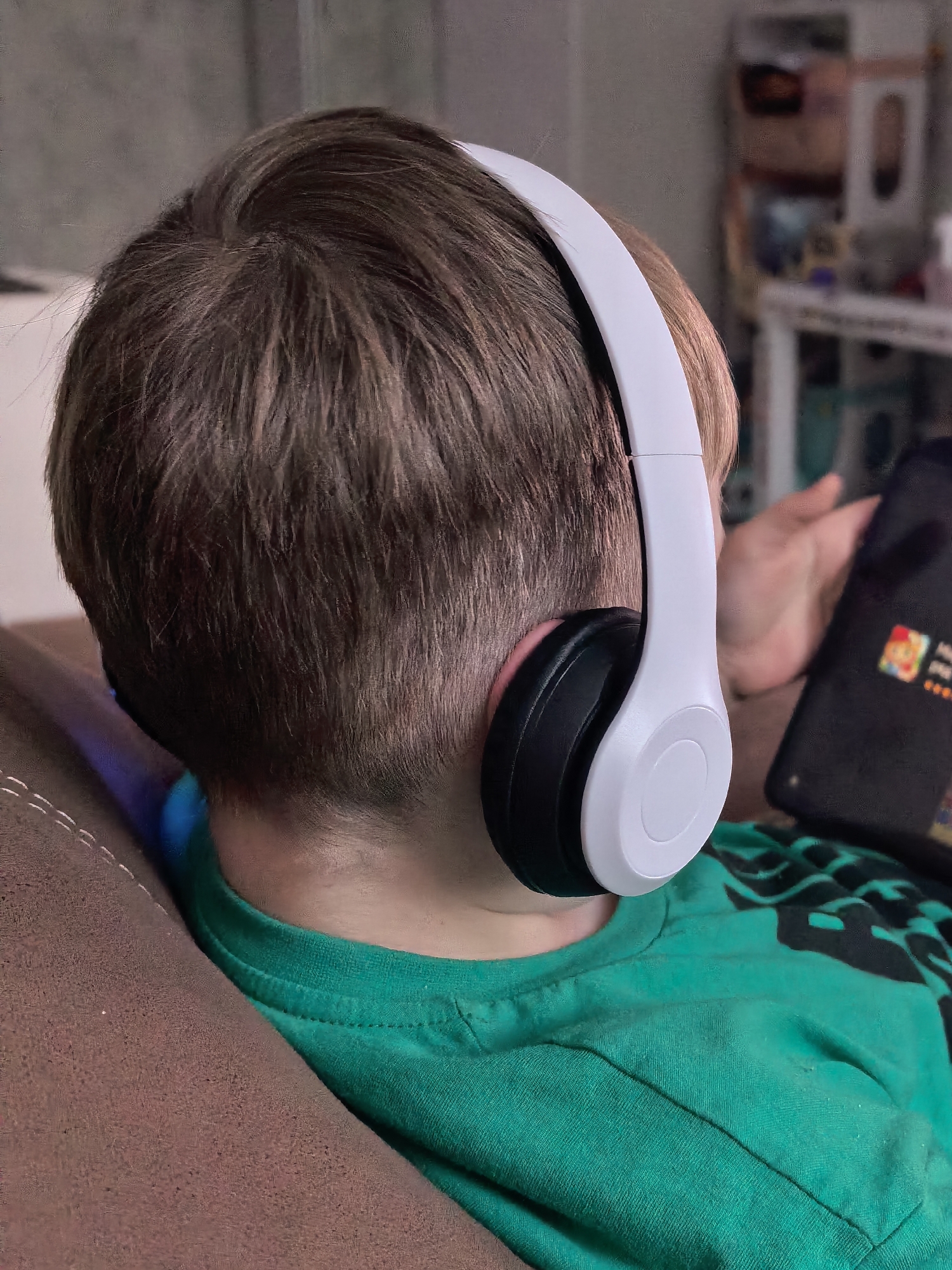 A young person is seen from behind, reclining on a brown upholstered piece of furniture. The individual is wearing a green t-shirt. They are wearing white headphones with black earcups. In their hand, they are holding a black mobile device displaying a game. In the background, there is a blurred white shelving unit with miscellaneous items. There is no information in the image to indicate a location in the city of Sterlitamak, Russia.
