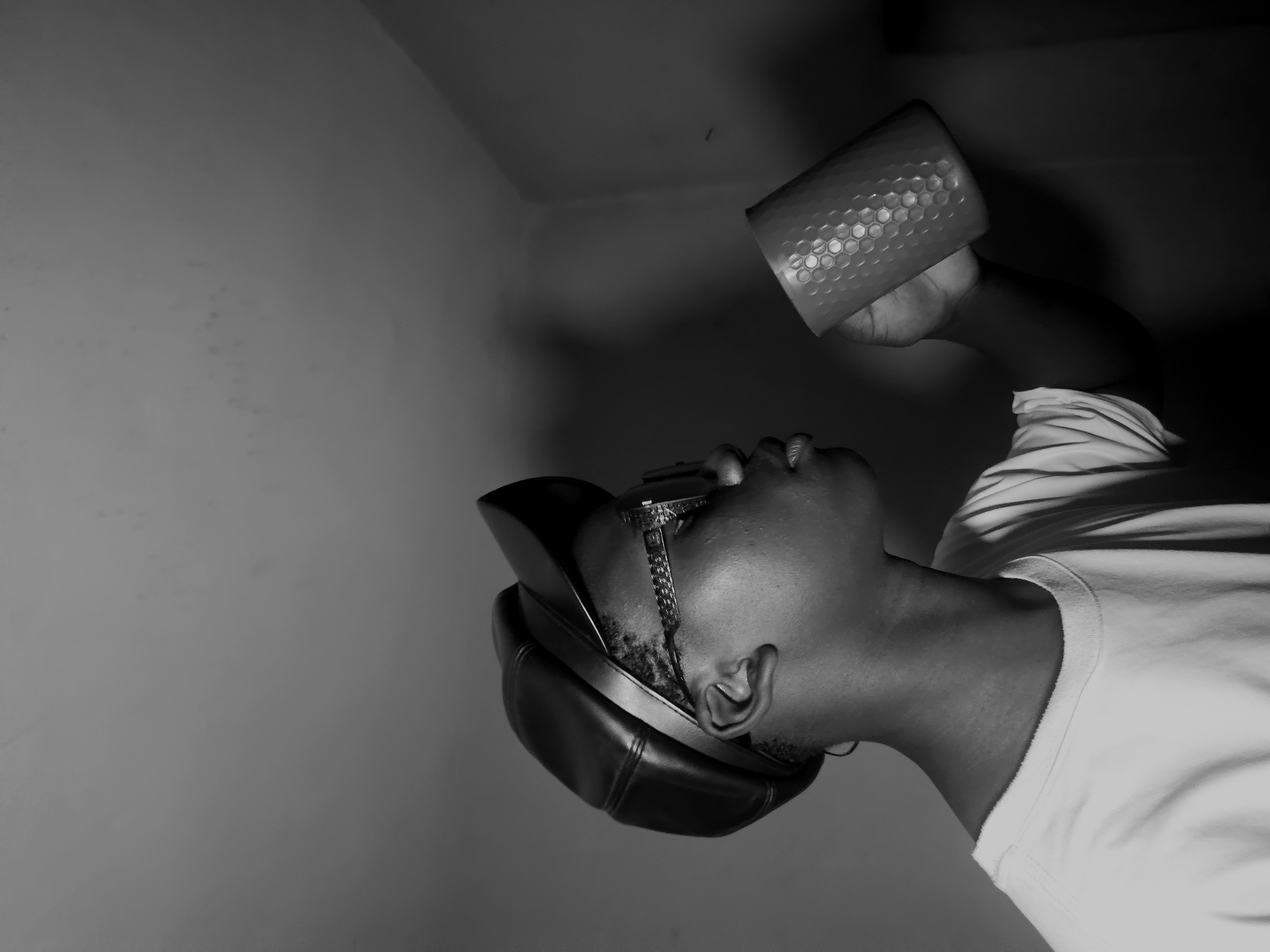 A black and white photo shows a dark-skinned man with a hat and glasses laying with his head tilted back in a room. He is wearing a white shirt. He is holding a honeycomb patterned cup over his head. The location in the city of Asaba, Nigeria cannot be determined.